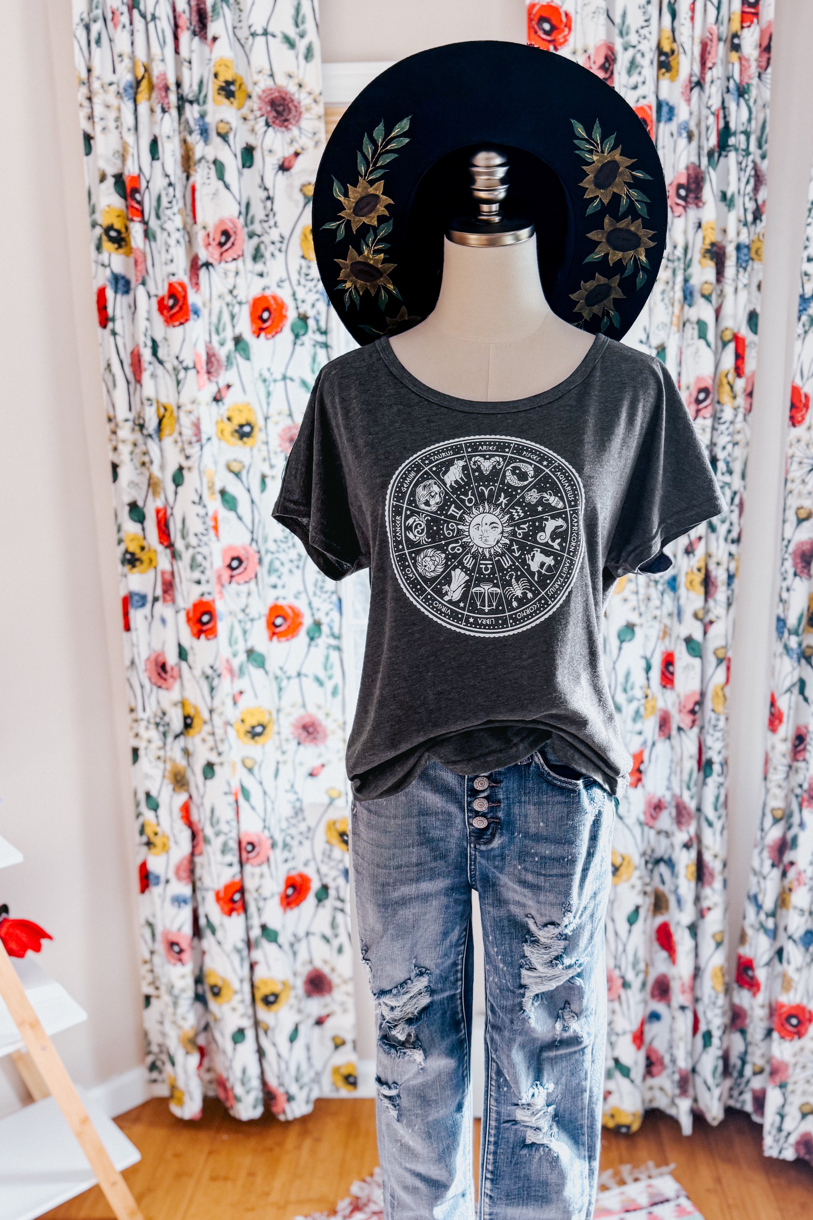 Mannequin wearing charcoal slouchy top with zodiac wheel print paired with distressed high-waist jeans and black hat with floral design indoors
