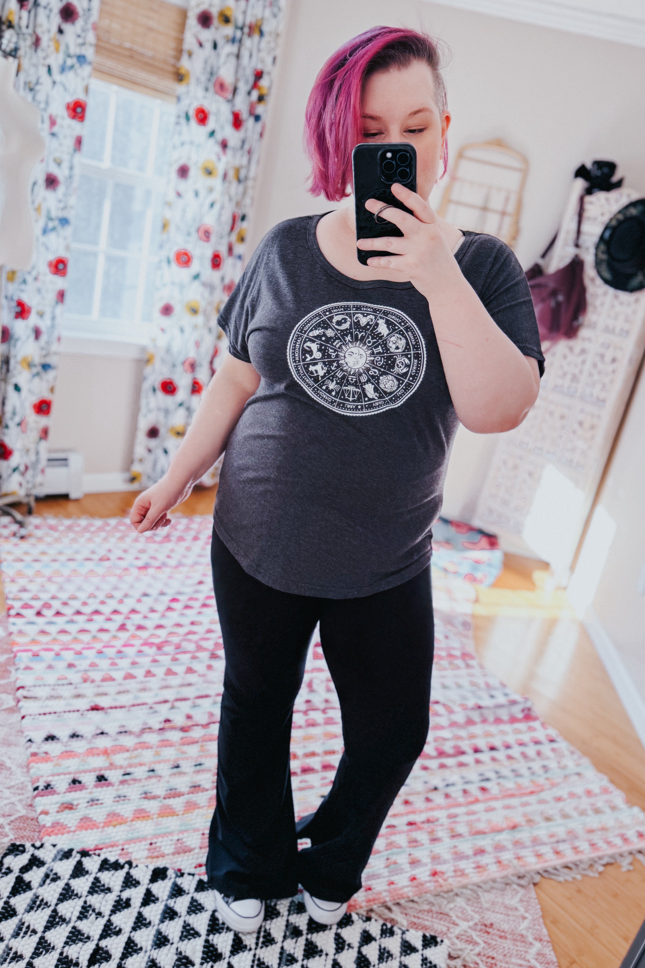 Woman with pink hair wearing charcoal slouchy top with zodiac design paired with black pants taking a selfie indoors
