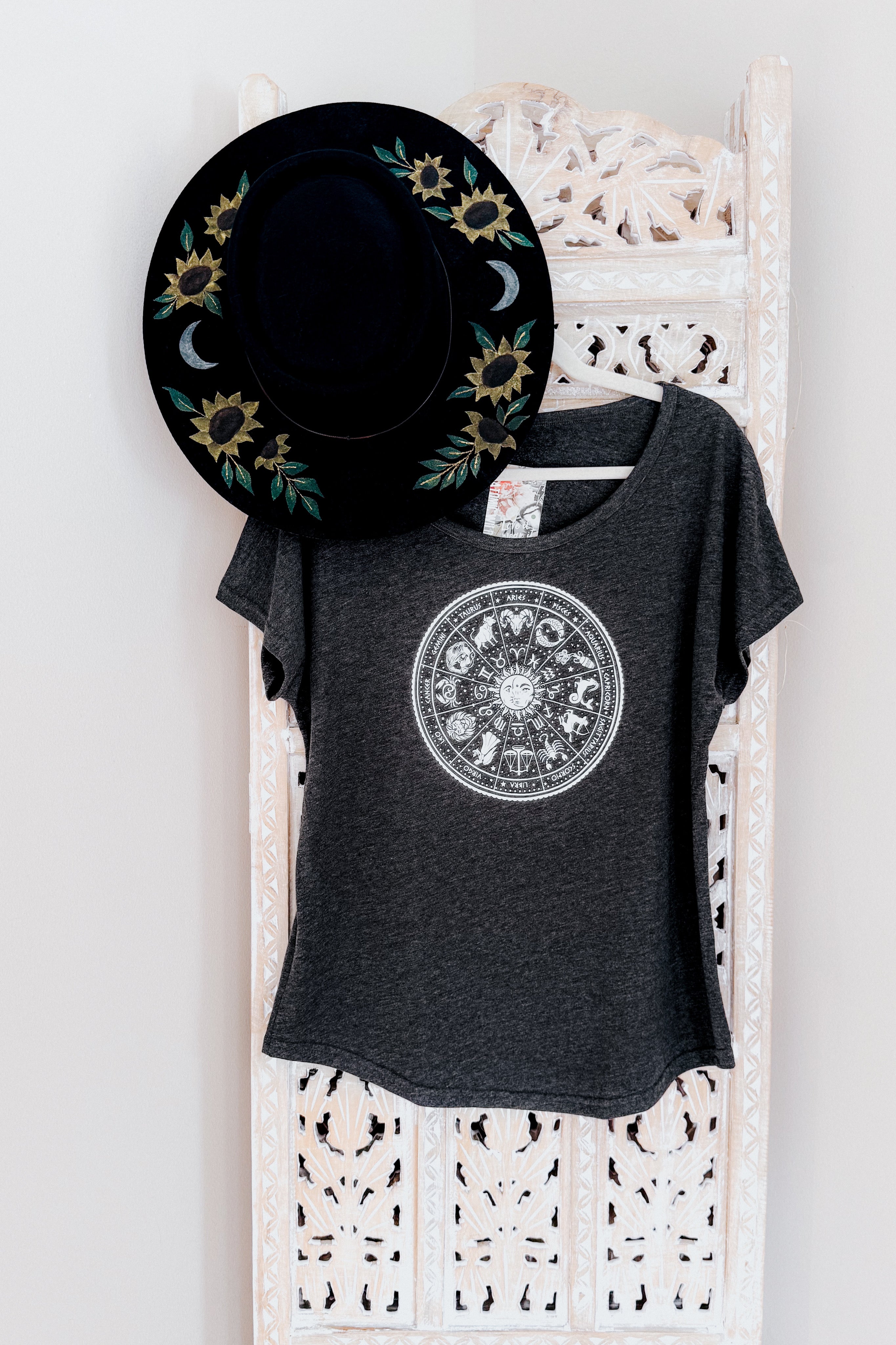Charcoal slouchy top with white zodiac wheel print paired with black hat featuring sunflowers and crescent moons hanging on carved screen