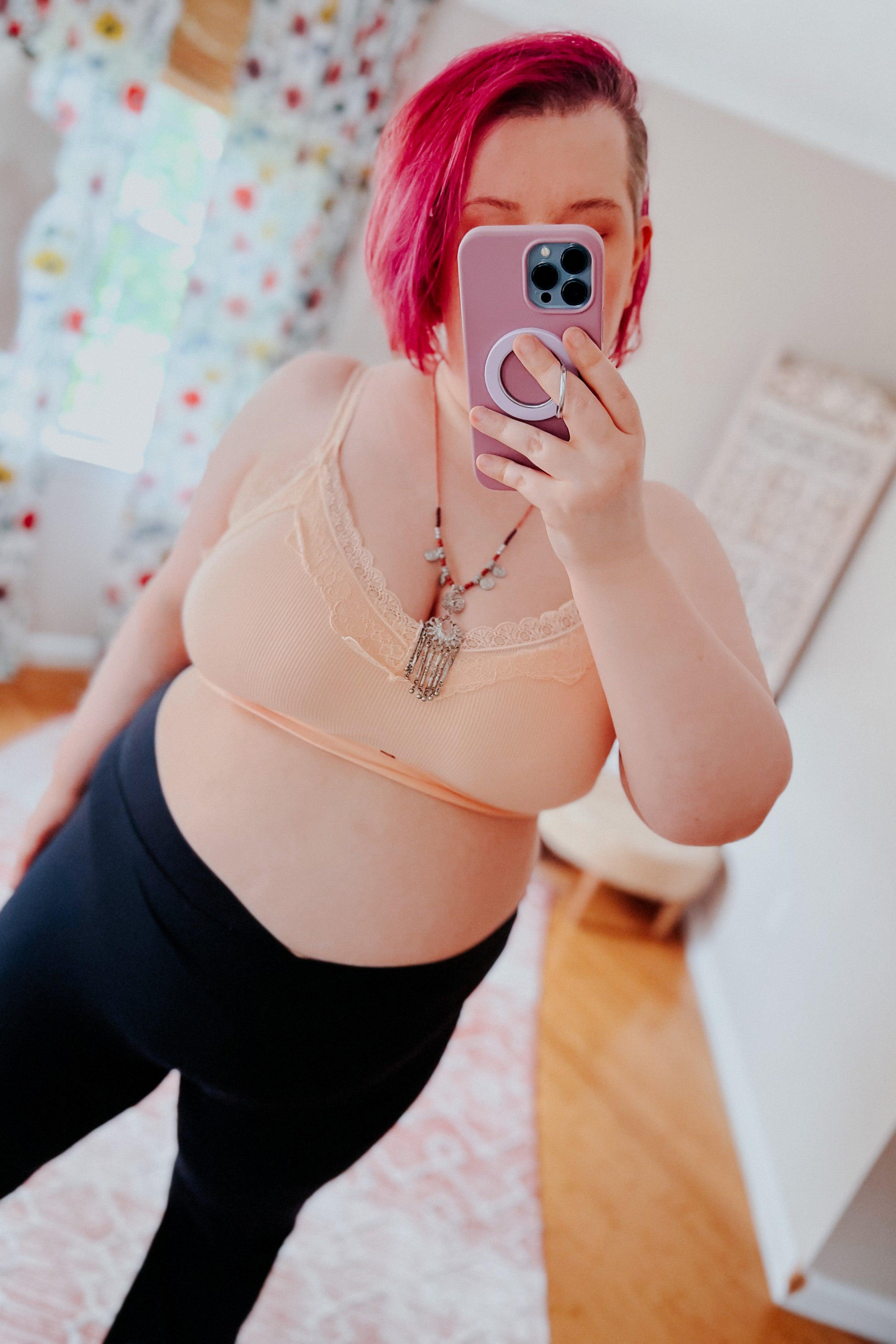 Plus-size woman with pink hair wearing peach lace trim bralette and black pants, taking selfie in softly lit room