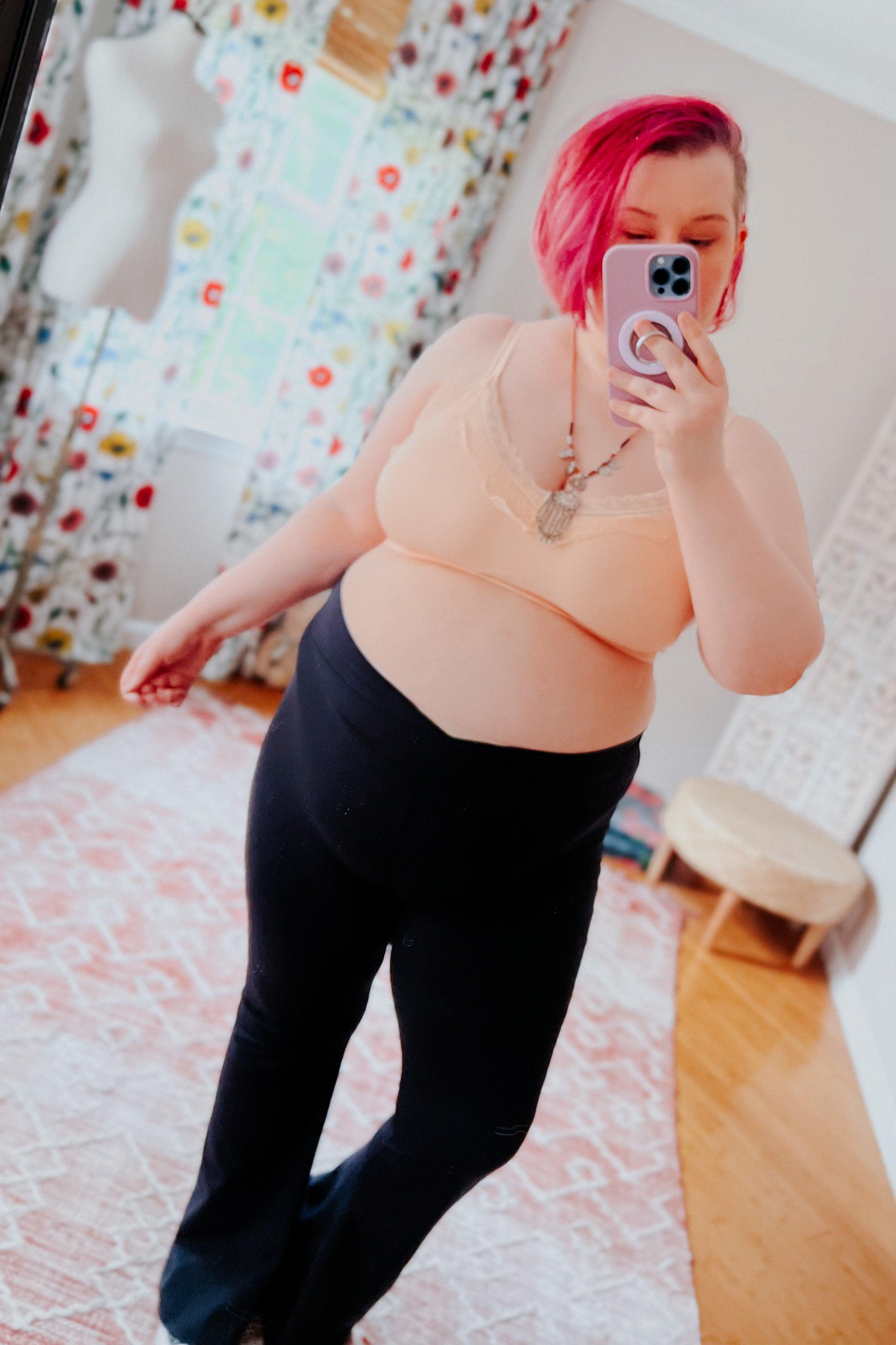 Plus-size woman with short pink hair wearing peach lace trim bralette and black pants taking mirror selfie in bright room