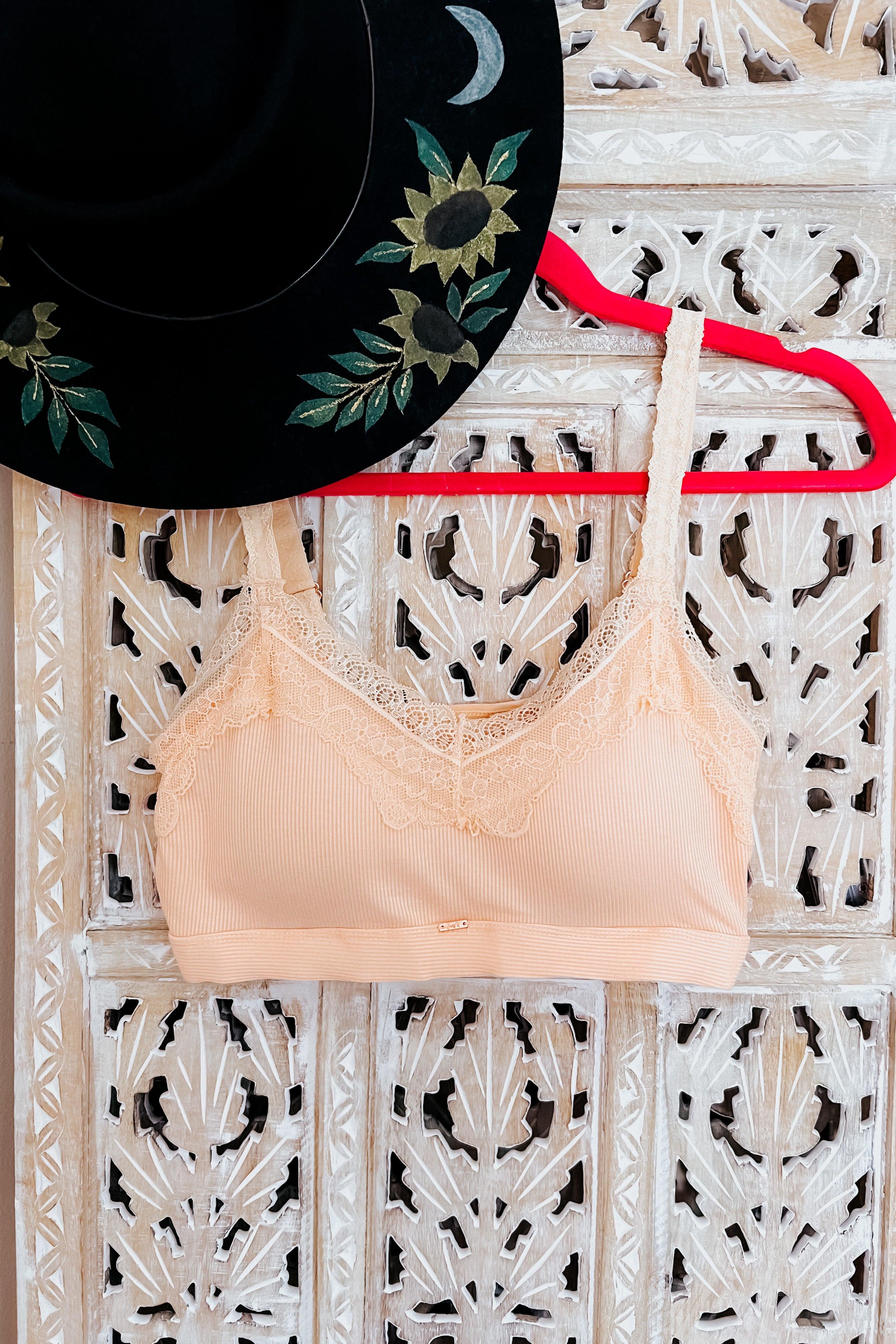 Peach bralette with lace trim hanging on red hanger beside black hat with floral and moon embroidery on carved wood panel