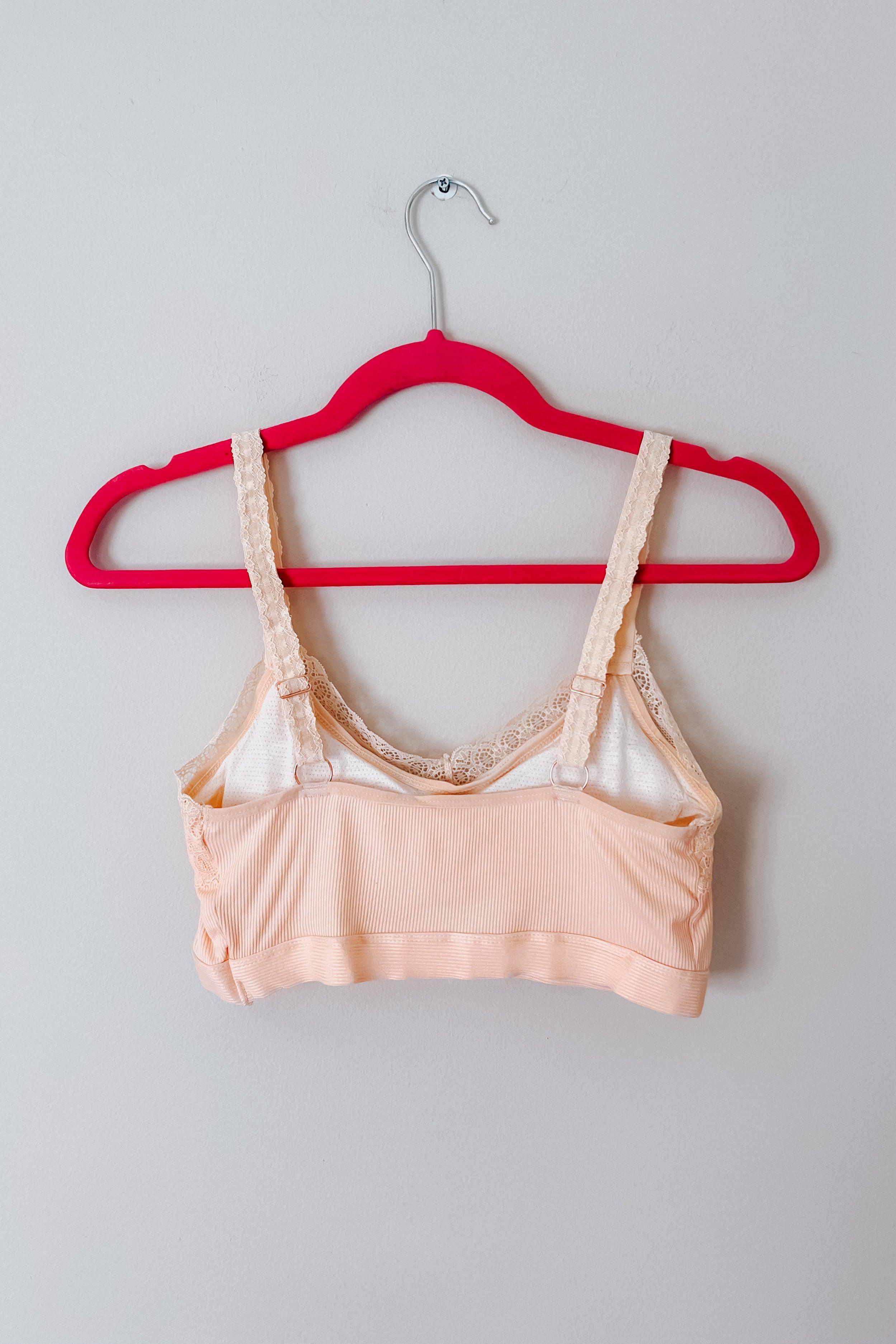 Peach lace-trim bralette with ribbed fabric and adjustable straps hanging on bright pink hanger against light gray wall