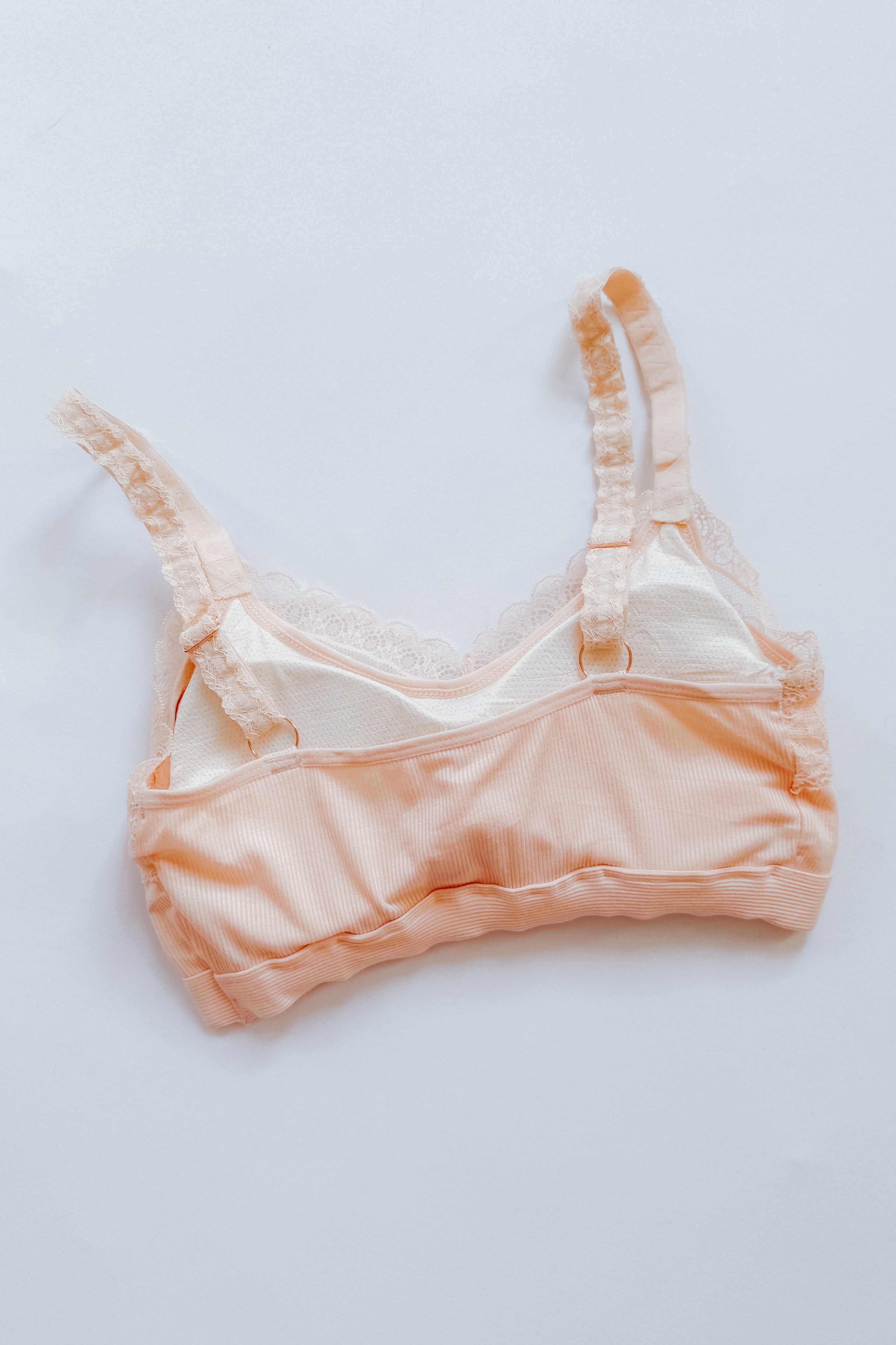 Peach-colored bralette with delicate lace trim and adjustable straps laid flat on a white background