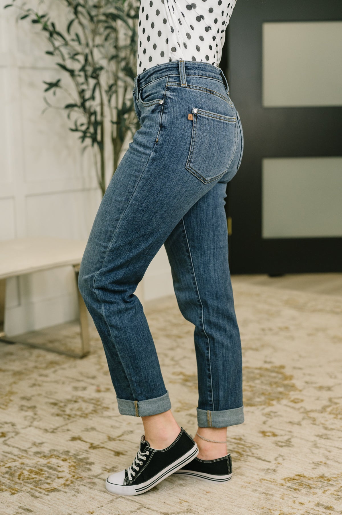 Woman modeling blue mid rise boyfriend jeans with cuffed hems and back pockets styled with a polka dot top and black sneakers indoors
