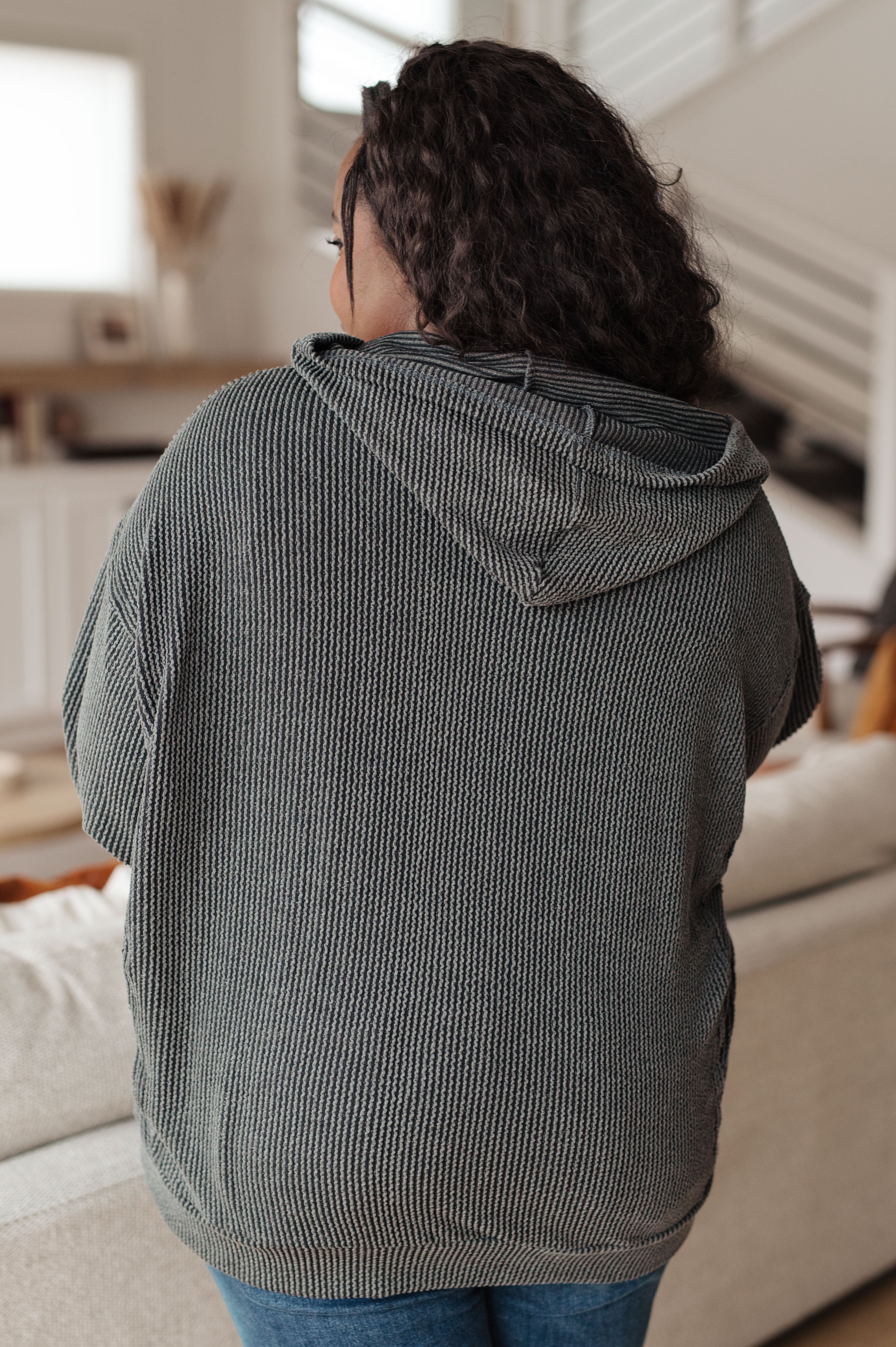 Woman with her back facing the camera wearing a grey rib knit hoodie over blue jeans against an indoor setting.