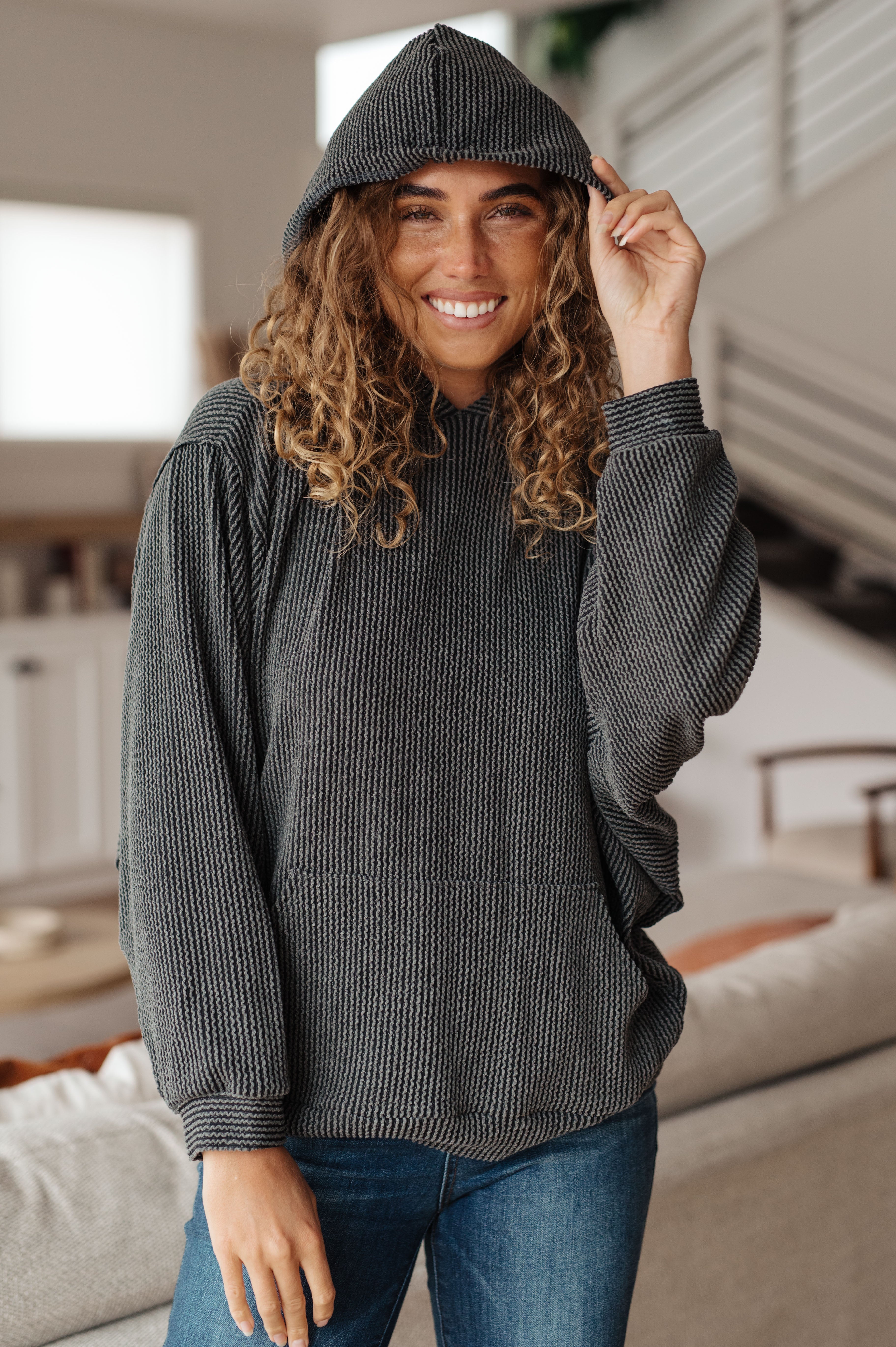 Woman wearing a grey rib knit hoodie over blue jeans against an indoor setting.