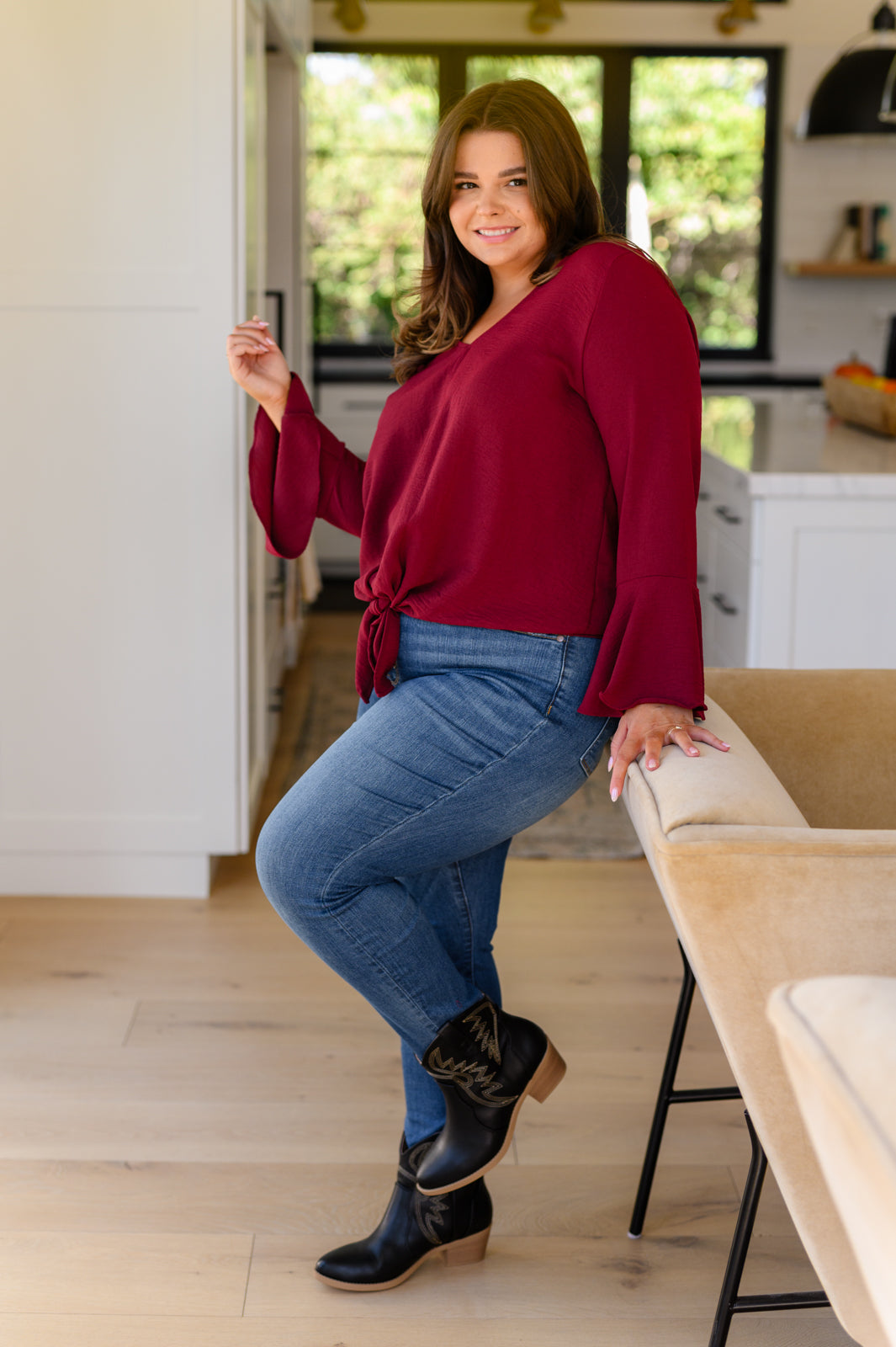 Smiling woman in bright kitchen wearing deep red flare sleeve top with front bow tied at waist and fitted blue jeans