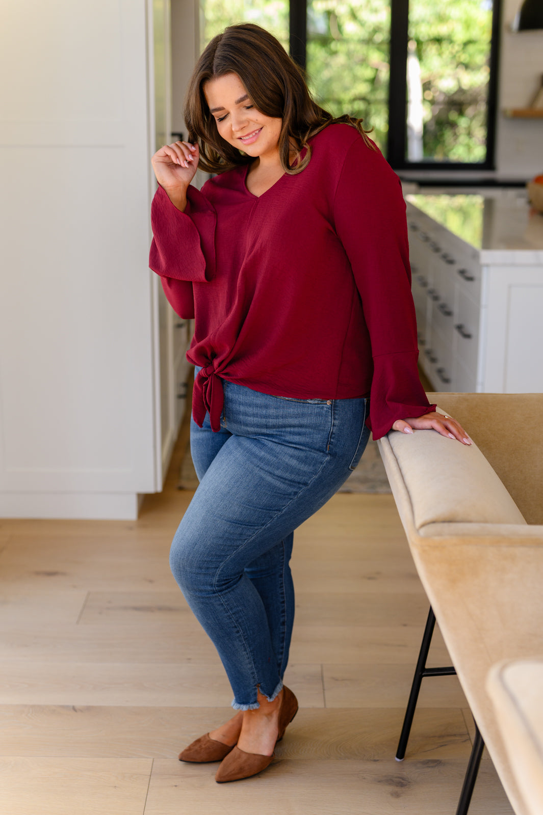 Curvy woman indoors wearing deep red V-neck top with front bow and flare sleeves paired with blue jeans and brown flats