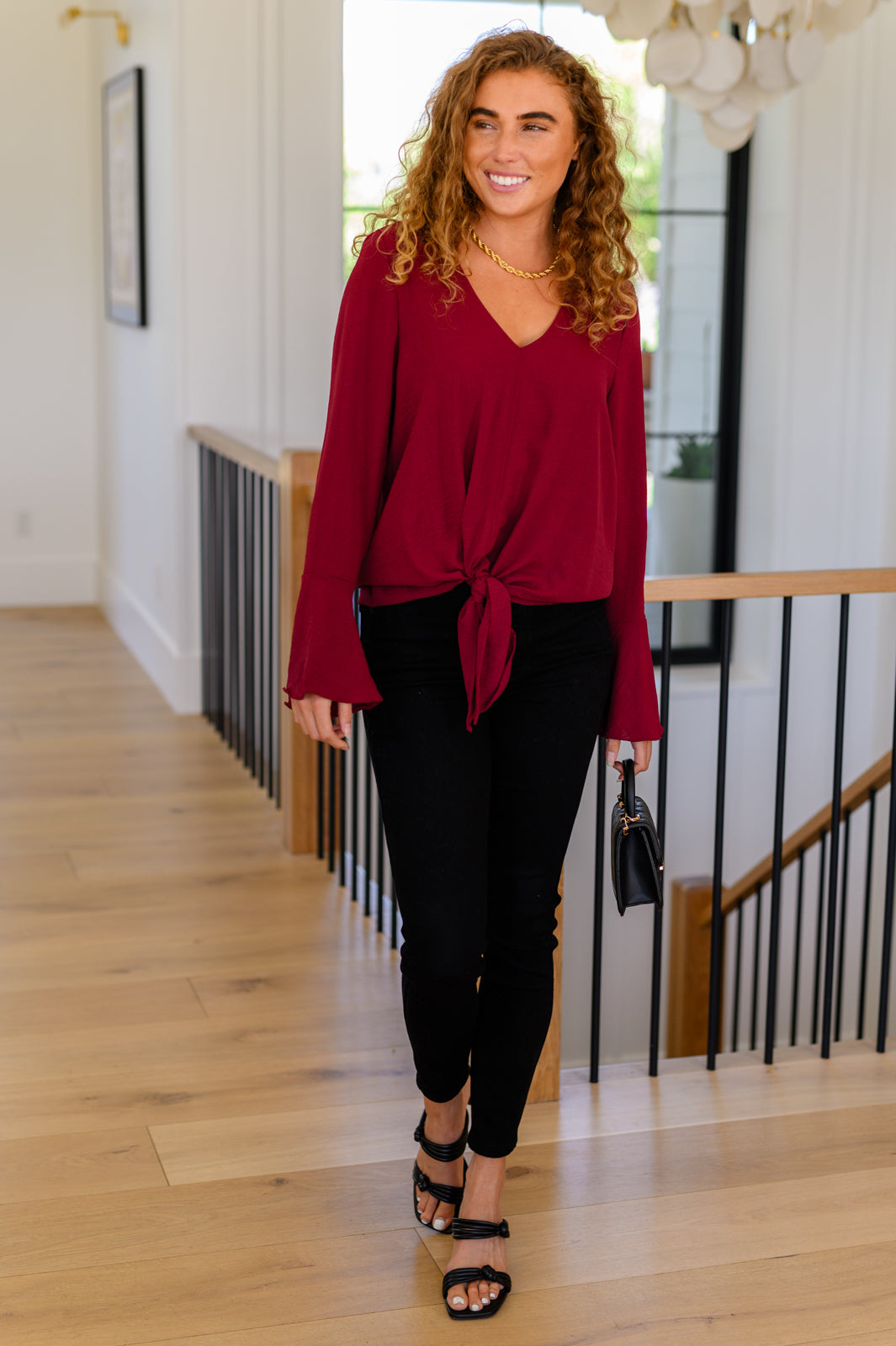 Smiling woman walking indoors wearing deep red V-neck top with front tie and flared sleeves holding black handbag