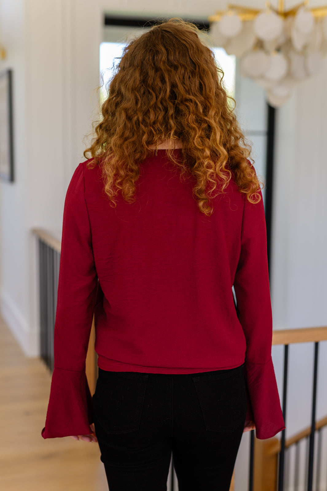 Woman with curly hair wearing deep red flare sleeve top standing indoors with back to camera near staircase railing