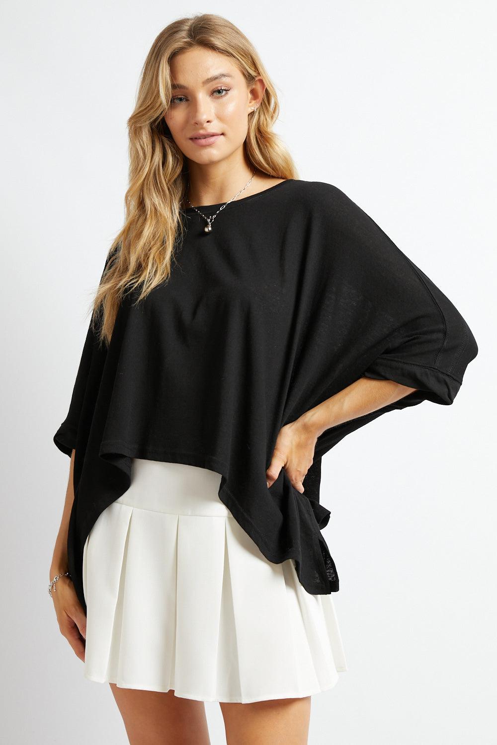 Blonde woman modeling loose black All My Life Boxy Top with draped sleeves paired with white pleated skirt against white background