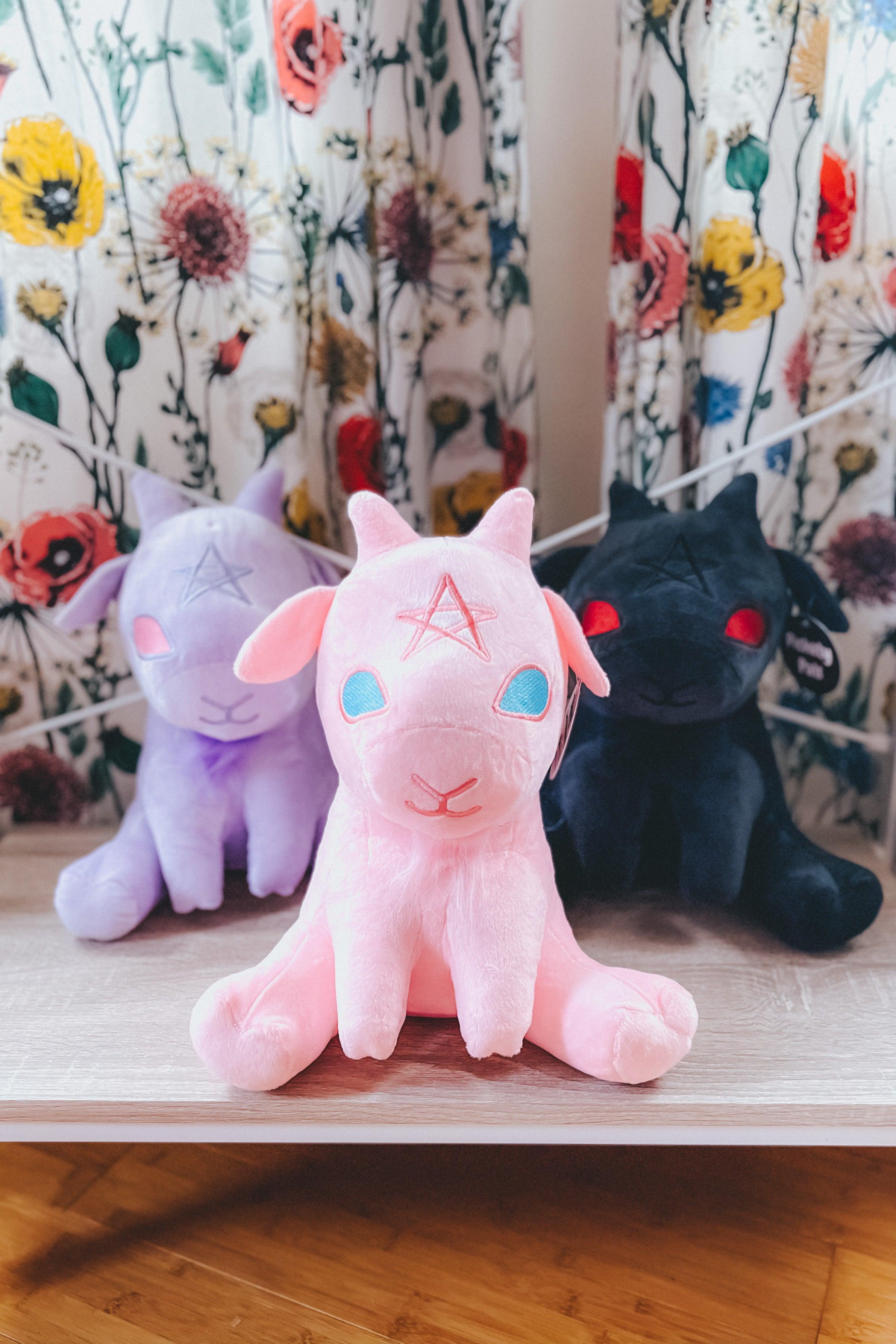 Cotton candy pink Baphy Witchy Baby Goat plush front and center with purple and black versions behind on wooden shelf