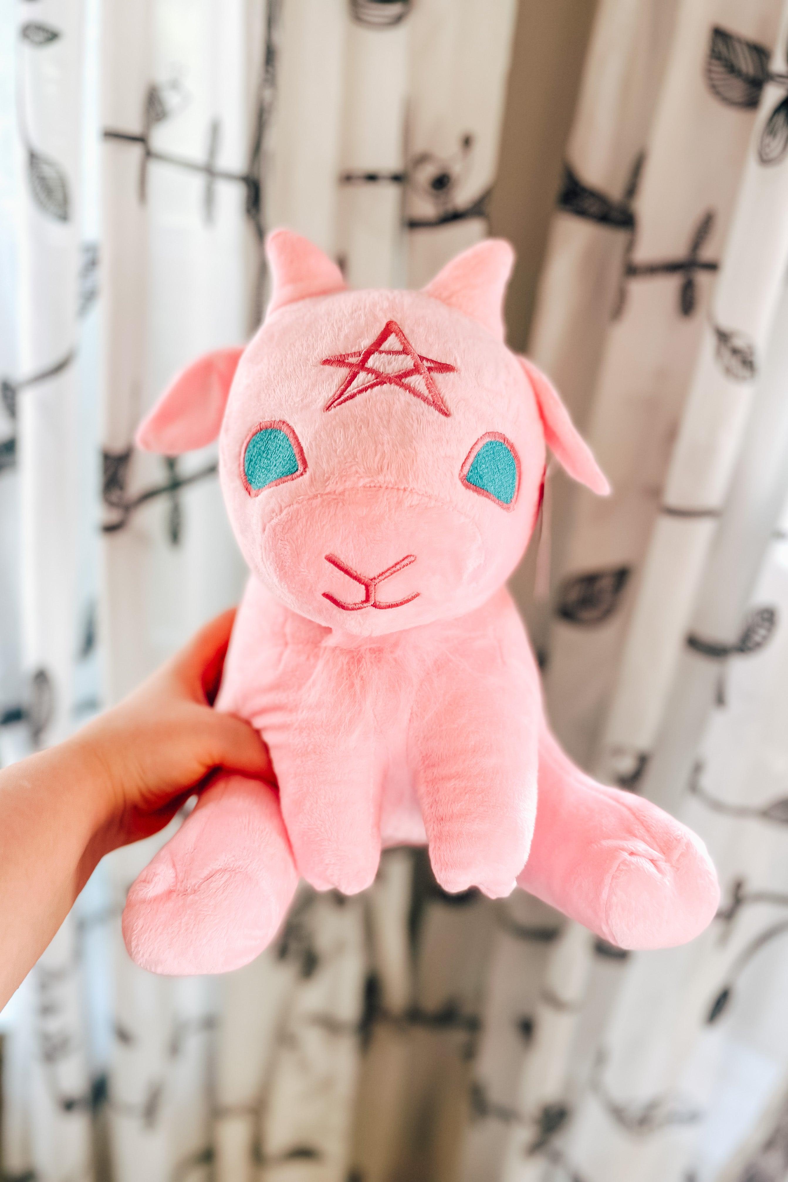 Hand holding cotton candy pink baby goat plush with star on forehead against white curtain with black leaf pattern