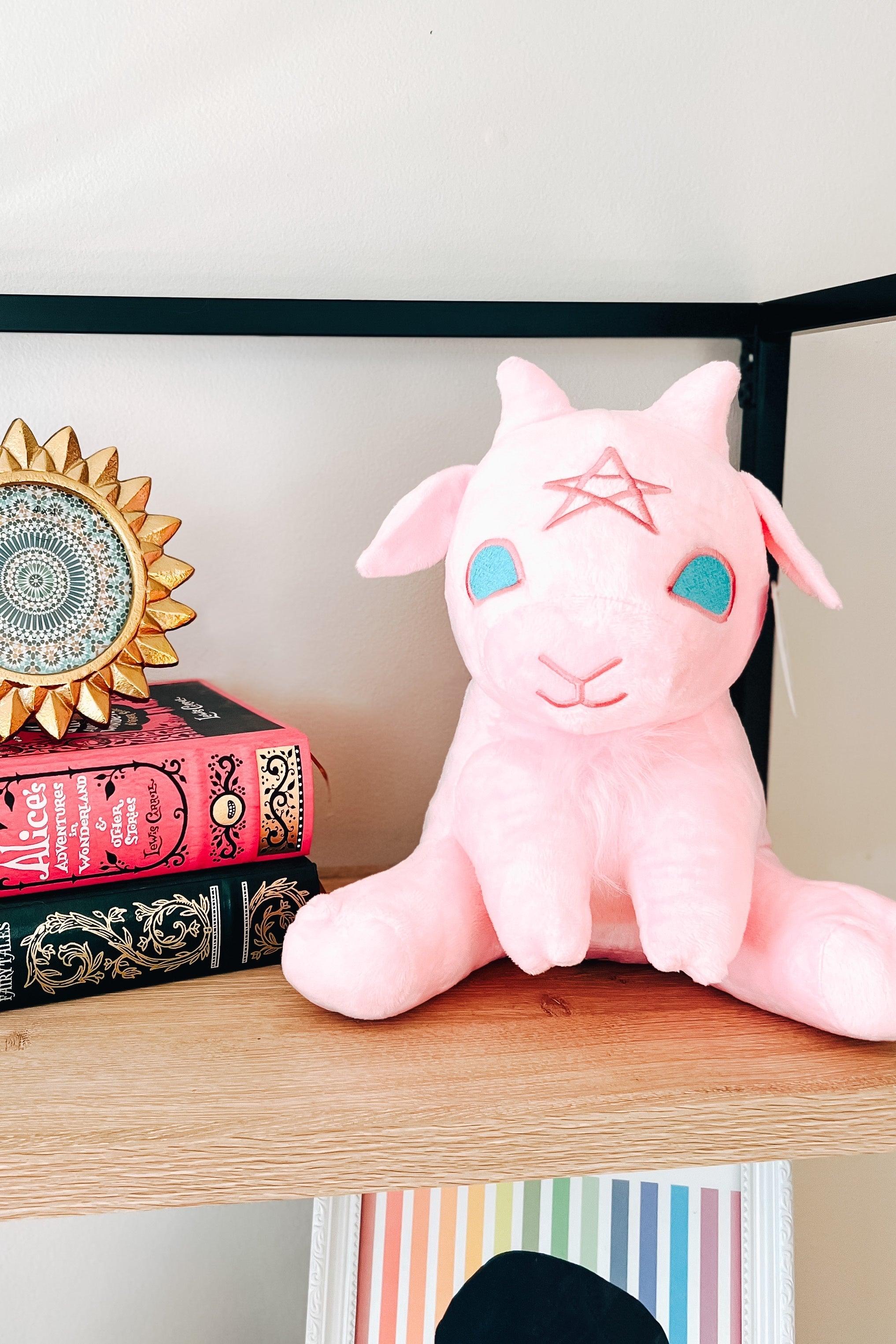 Cotton candy pink witchy baby goat plush with blue eyes and star on forehead seated on wooden shelf next to decorative books