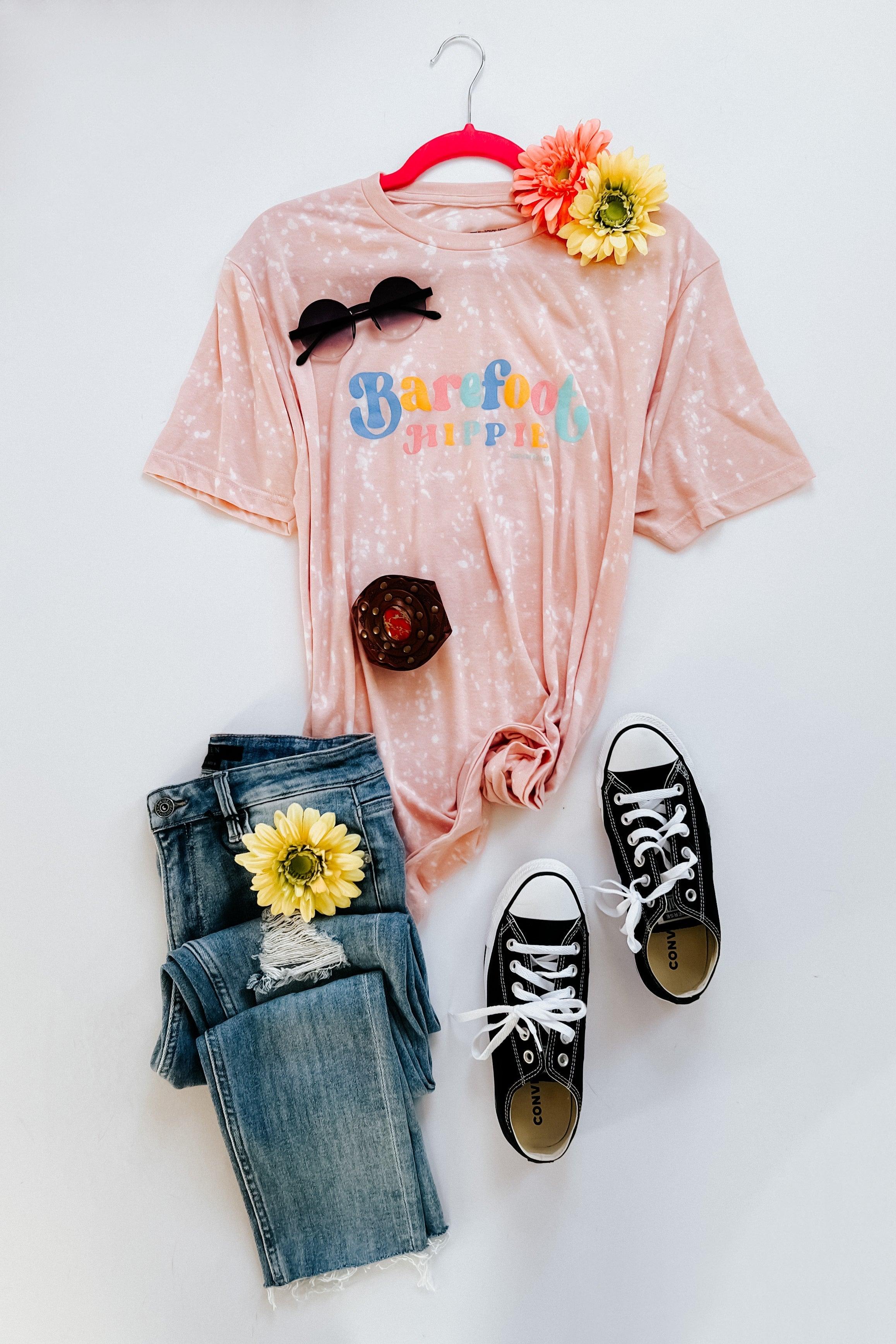 Pink bleached Barefoot Hippie tee styled with denim jeans, black sneakers, sunglasses, and floral accents on white background