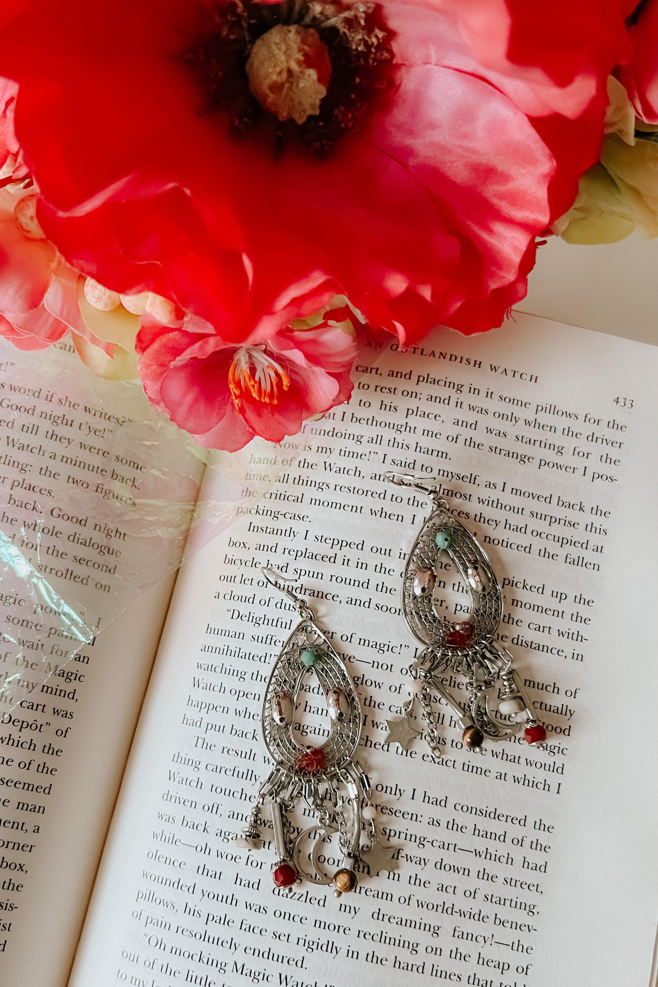 Delicate silver teardrop earrings with dangling beads and charms, placed on an open book beneath bright red flowers