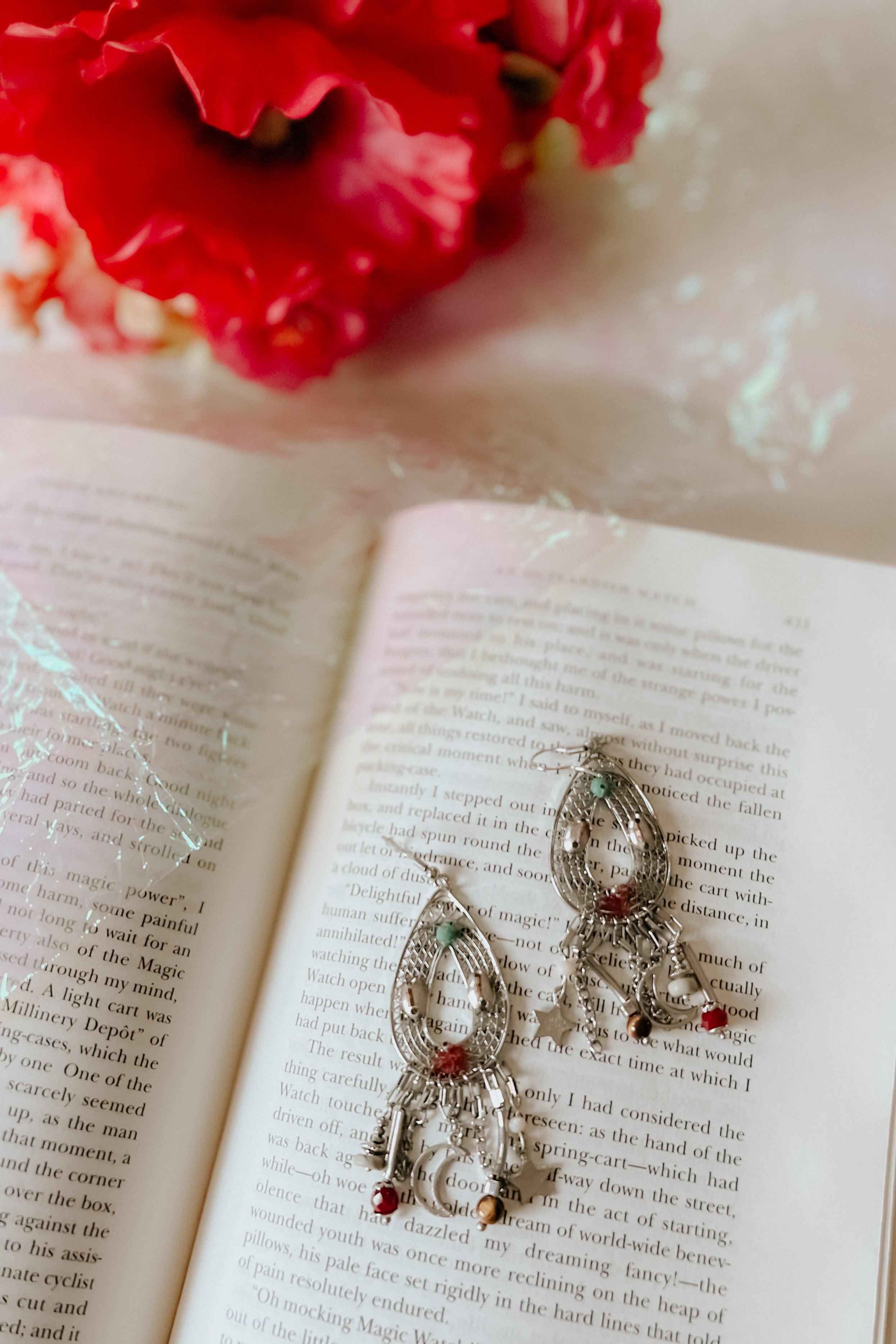 Silver teardrop earrings with moon and star charms and red and turquoise beads displayed on an open book page with flowers nearby
