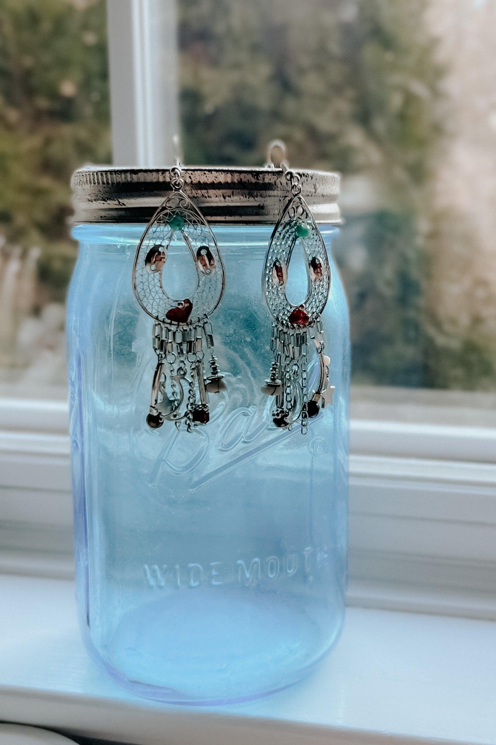 Intricate silver teardrop earrings with dangling beads and charms hanging on a blue glass jar lid by a window