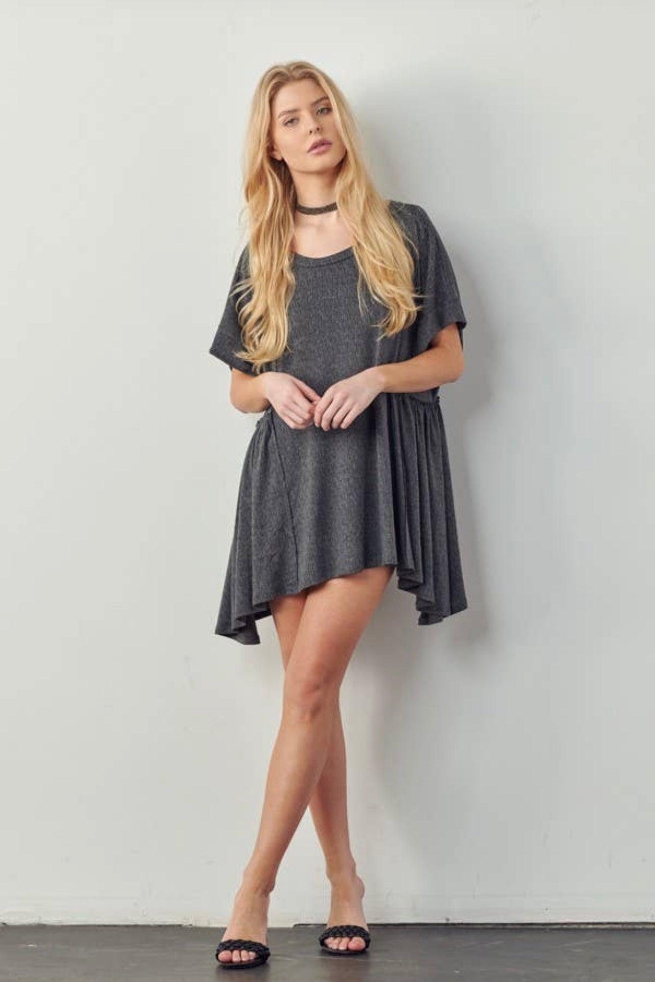 Woman with long blonde hair wearing loose dark gray asymmetrical hem tunic with short sleeves and black sandals standing against wall