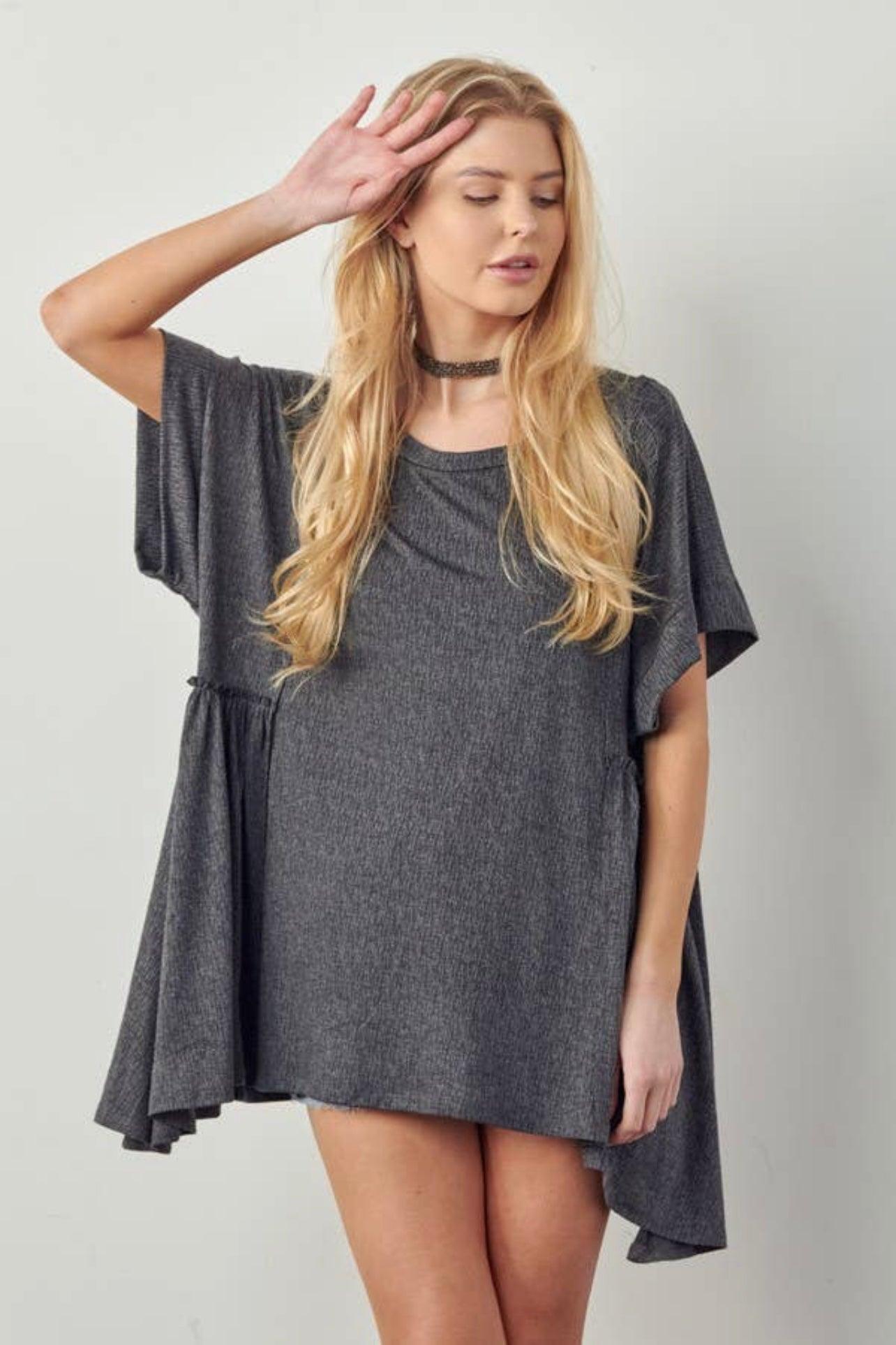 Blonde woman wearing loose charcoal gray oversized tunic with shark bite hem and short sleeves standing against plain background