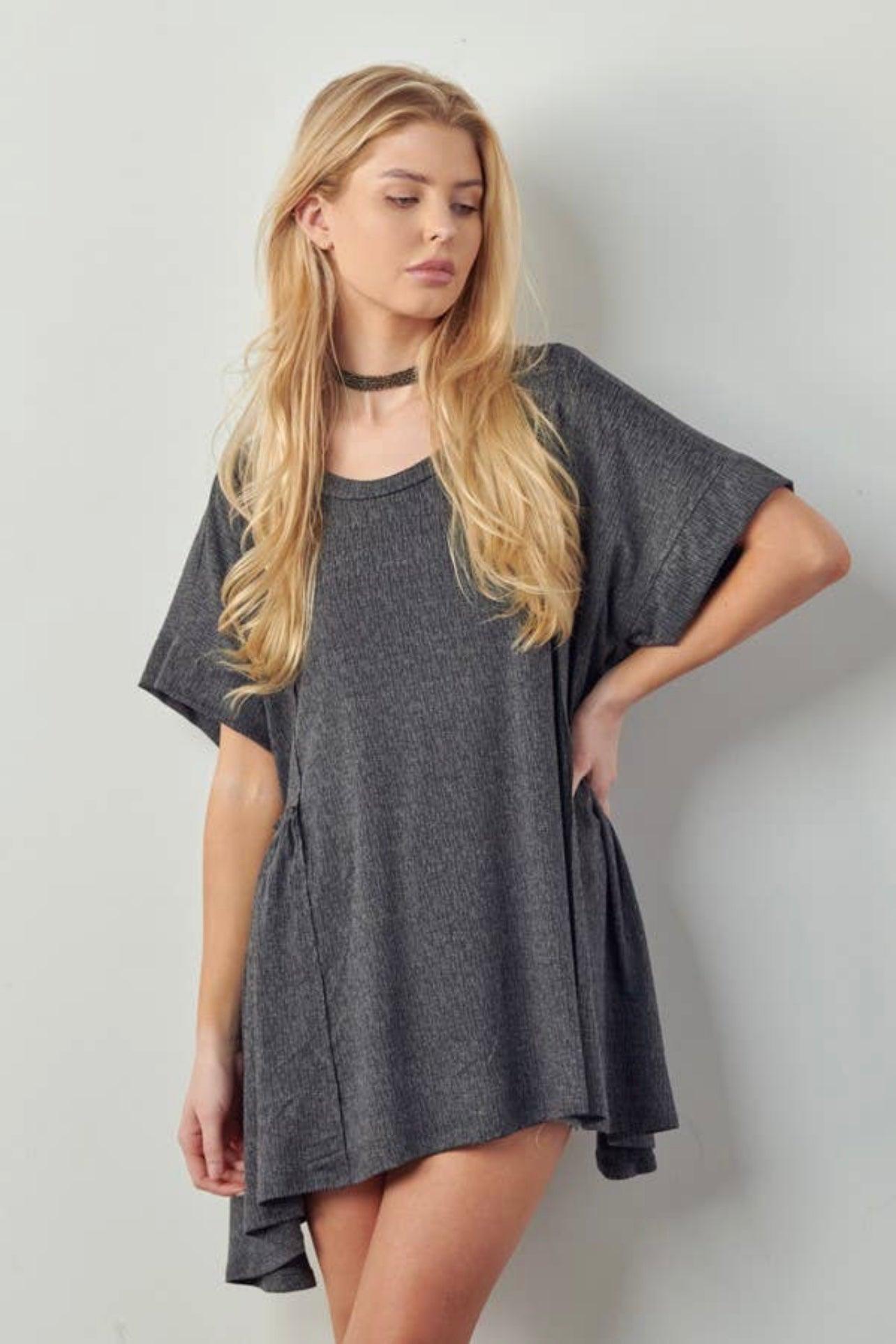 Blonde woman wearing loose dark gray oversized tunic with short sleeves standing against light wall