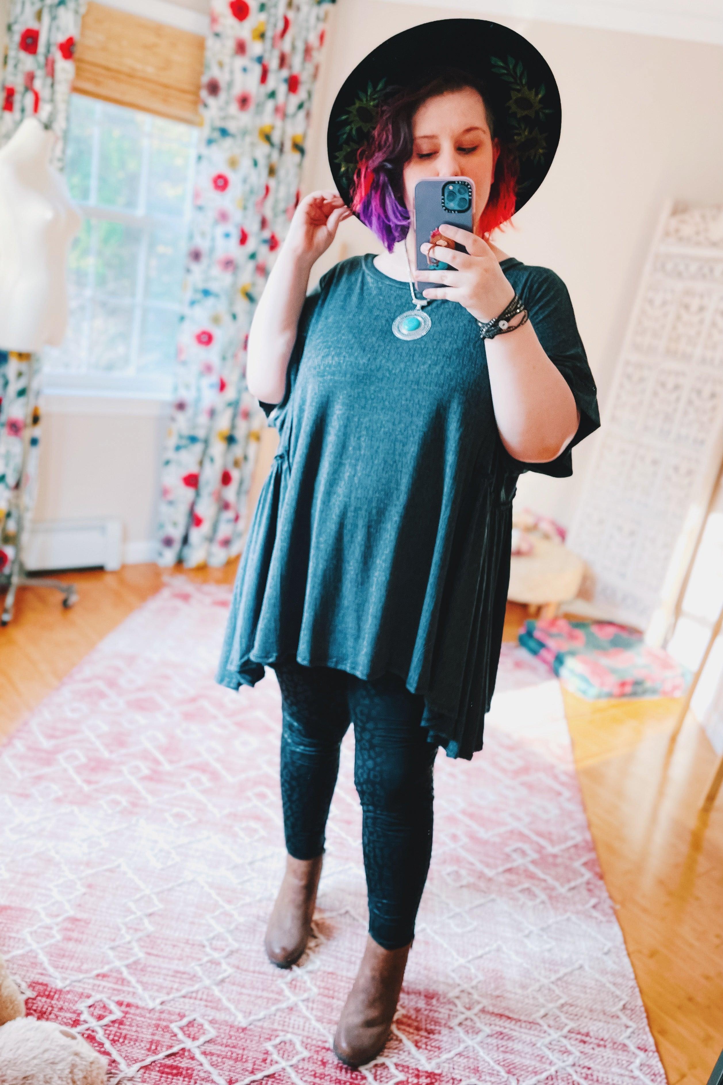 Woman taking mirror selfie wearing oversized dark tunic with black hat, patterned leggings, and ankle boots in bright room