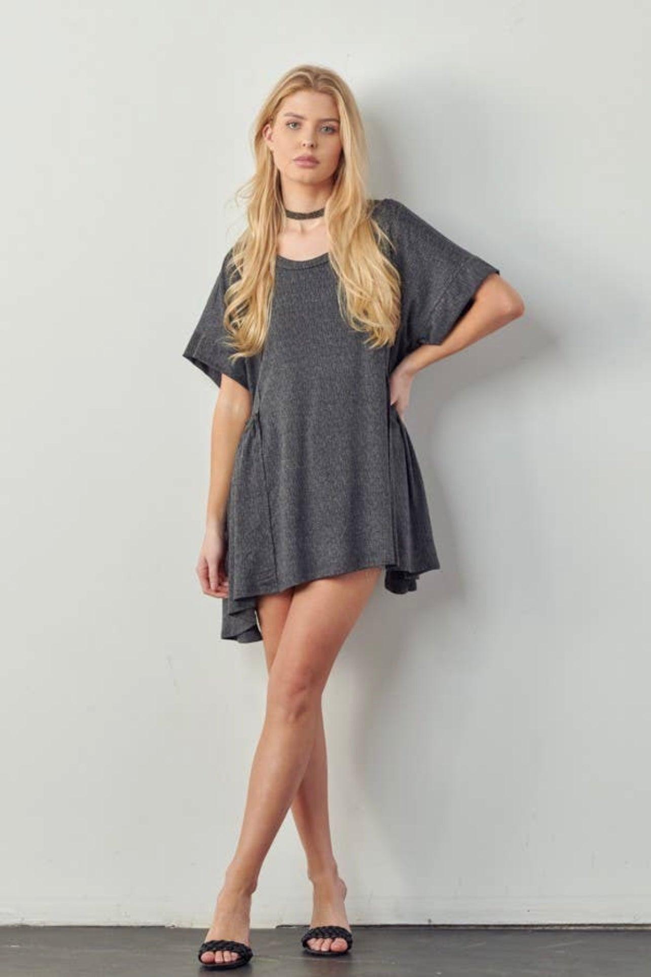Blonde woman wearing loose dark gray oversized tunic dress with short sleeves and black sandals indoors