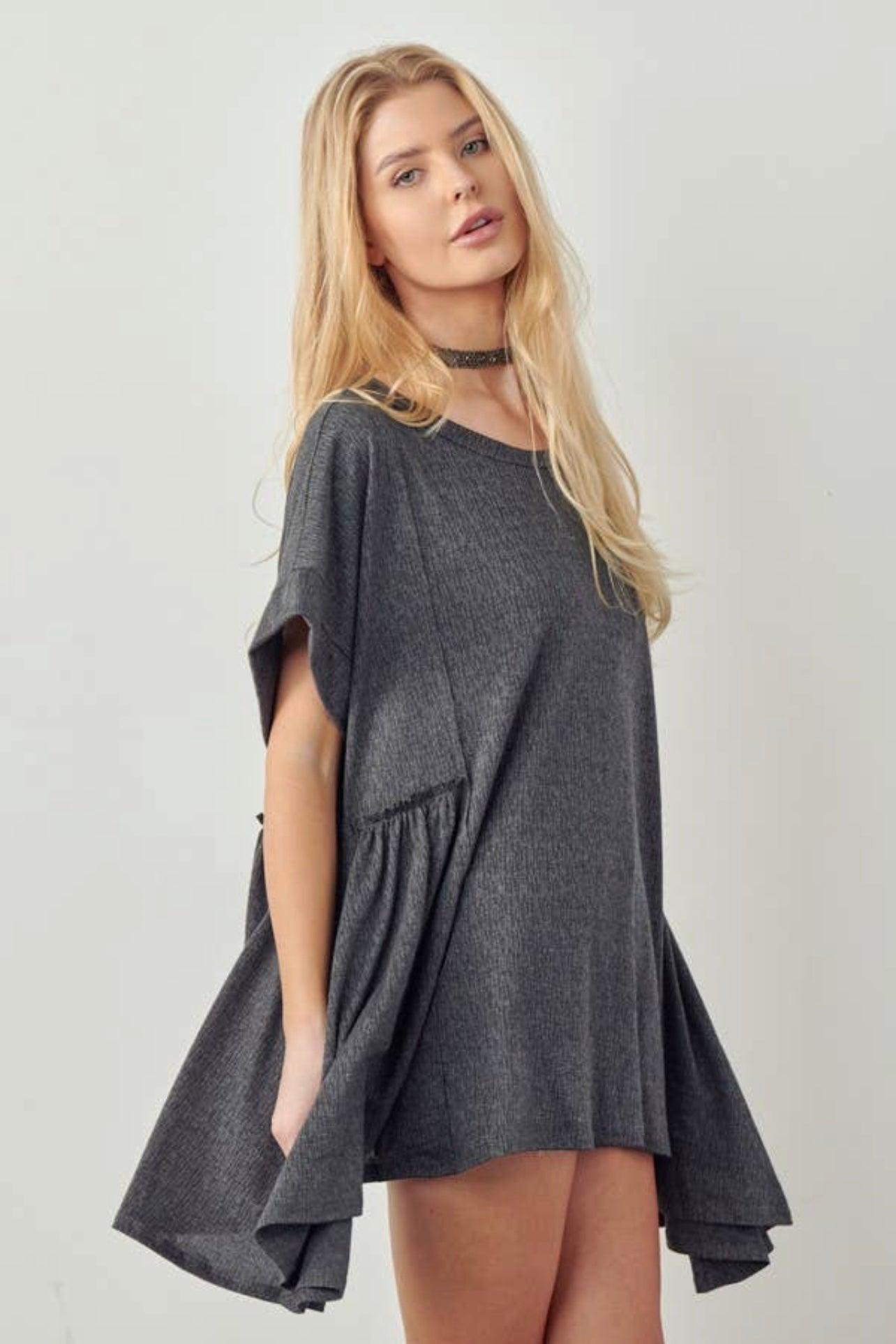 Blonde woman wearing loose dark gray oversized tunic with short sleeves and flared sides against plain background
