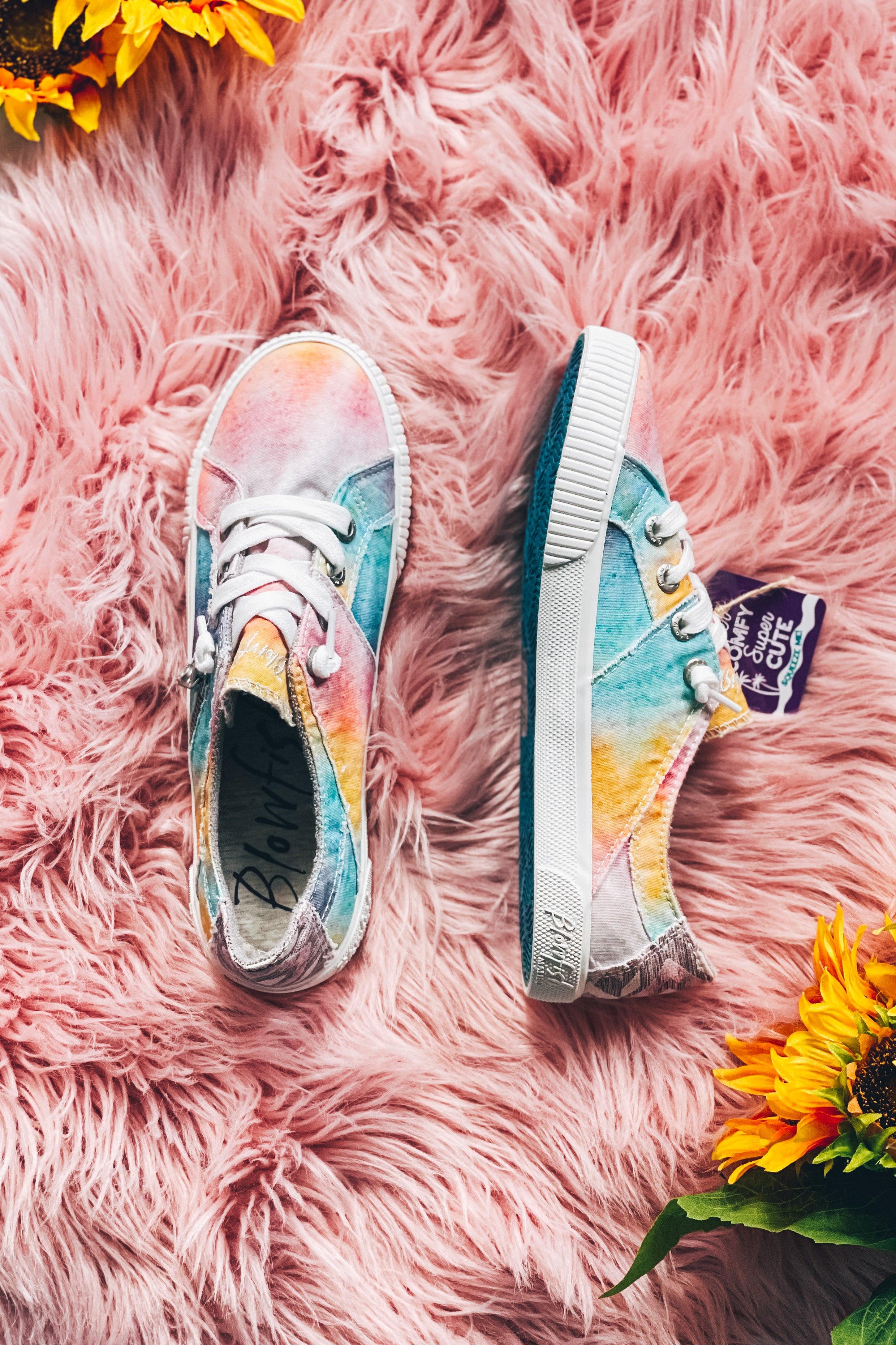 Pair of pastel tie-dye sneakers with white laces on soft pink furry surface next to bright sunflowers
