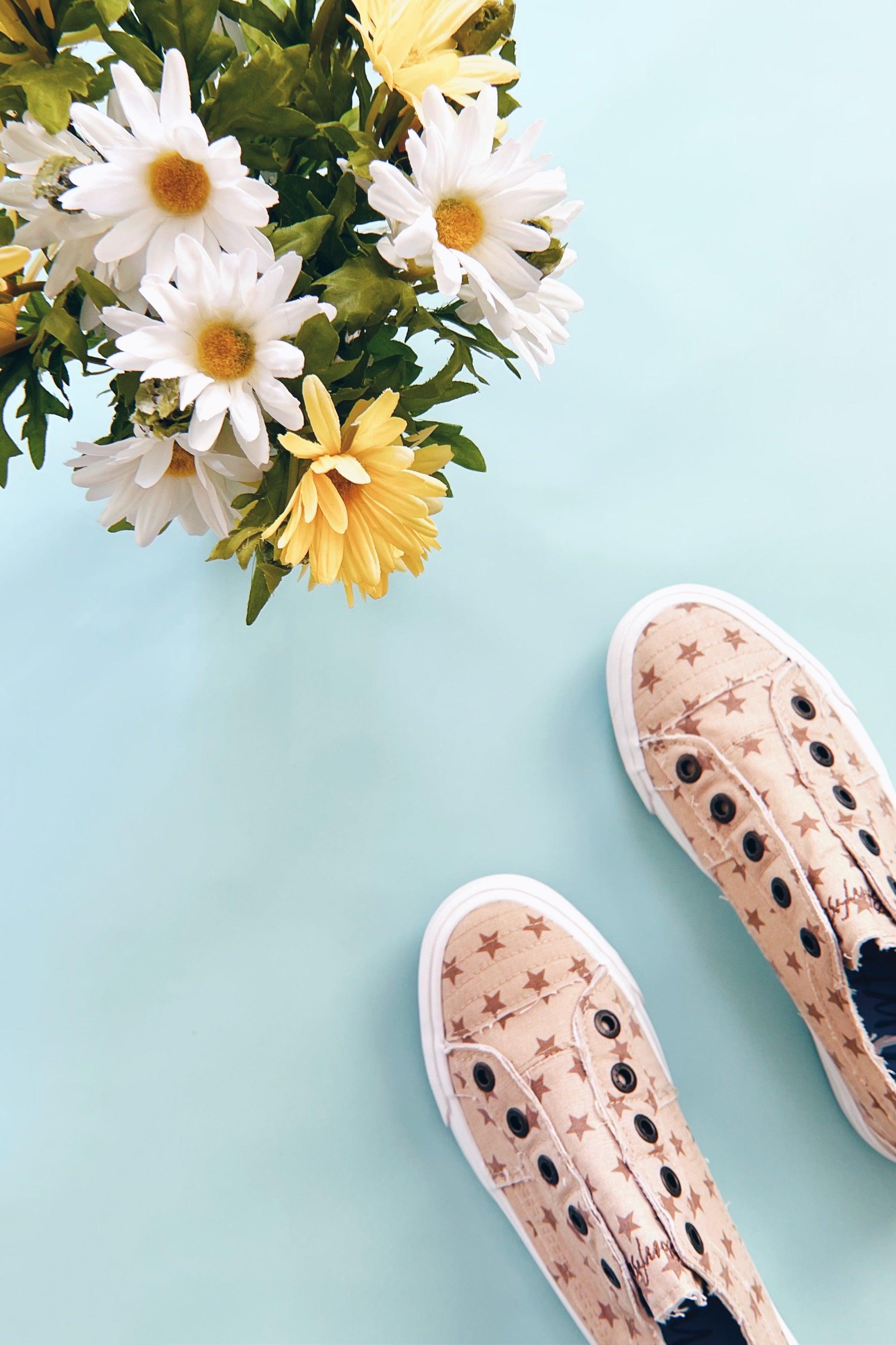 Natural beige canvas slip-on sneakers with brown star pattern on light blue background next to yellow and white daisies