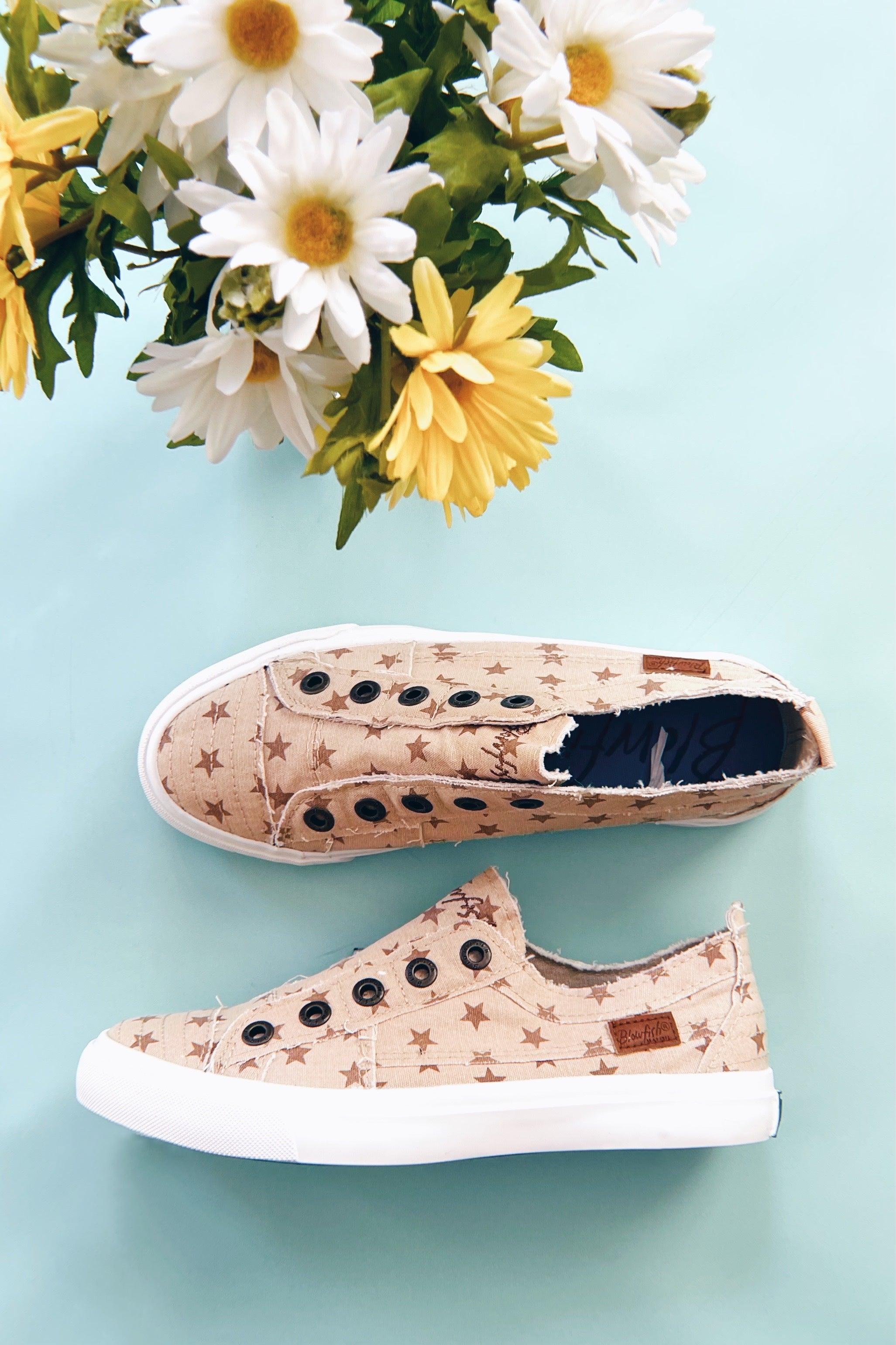 Beige canvas slip-on sneakers with star print and black eyelets displayed next to white and yellow daisies on blue background