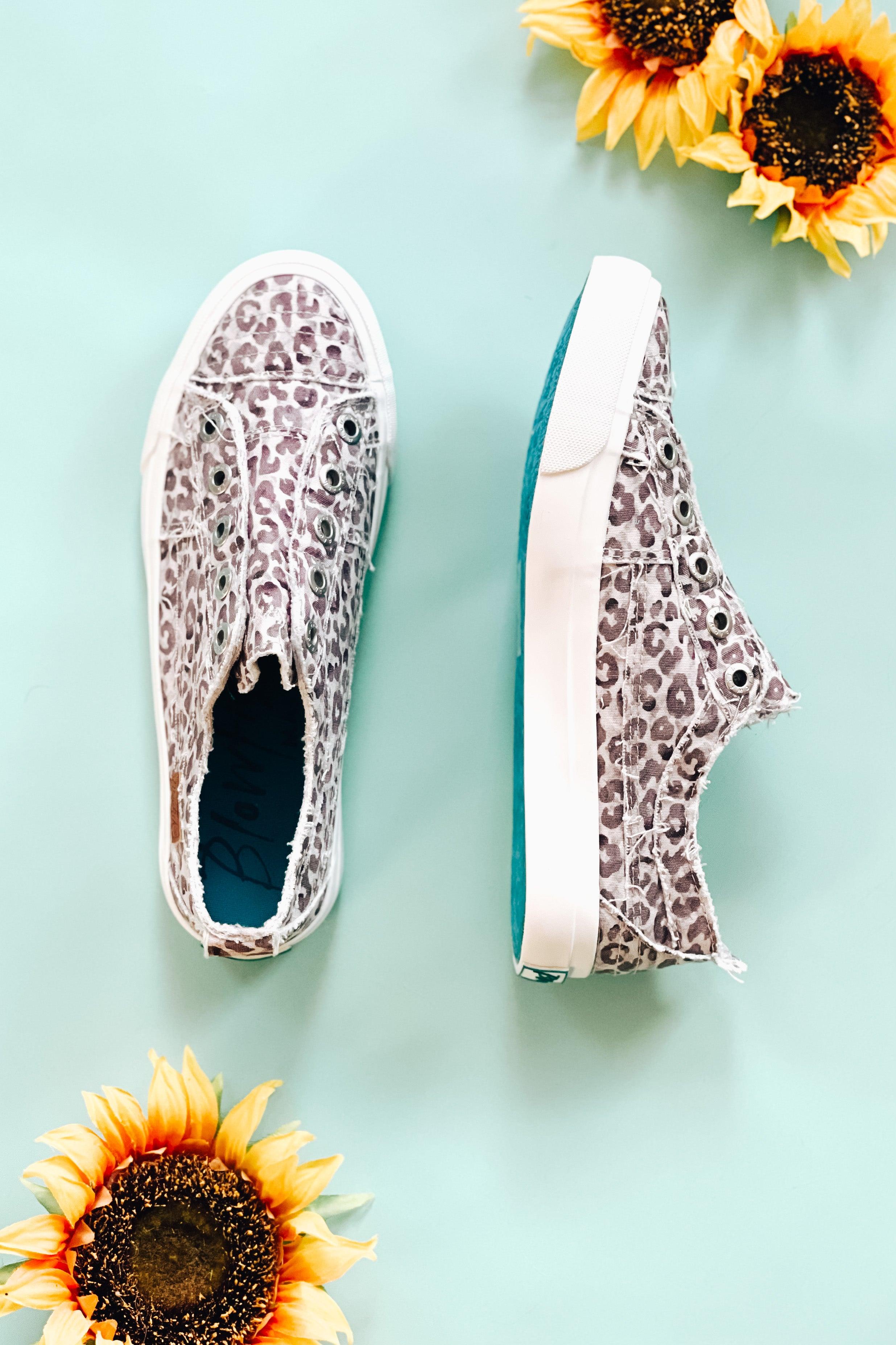 Frayed purple gray water leopard slip-on sneakers with teal sole on pale blue surface with two sunflowers nearby