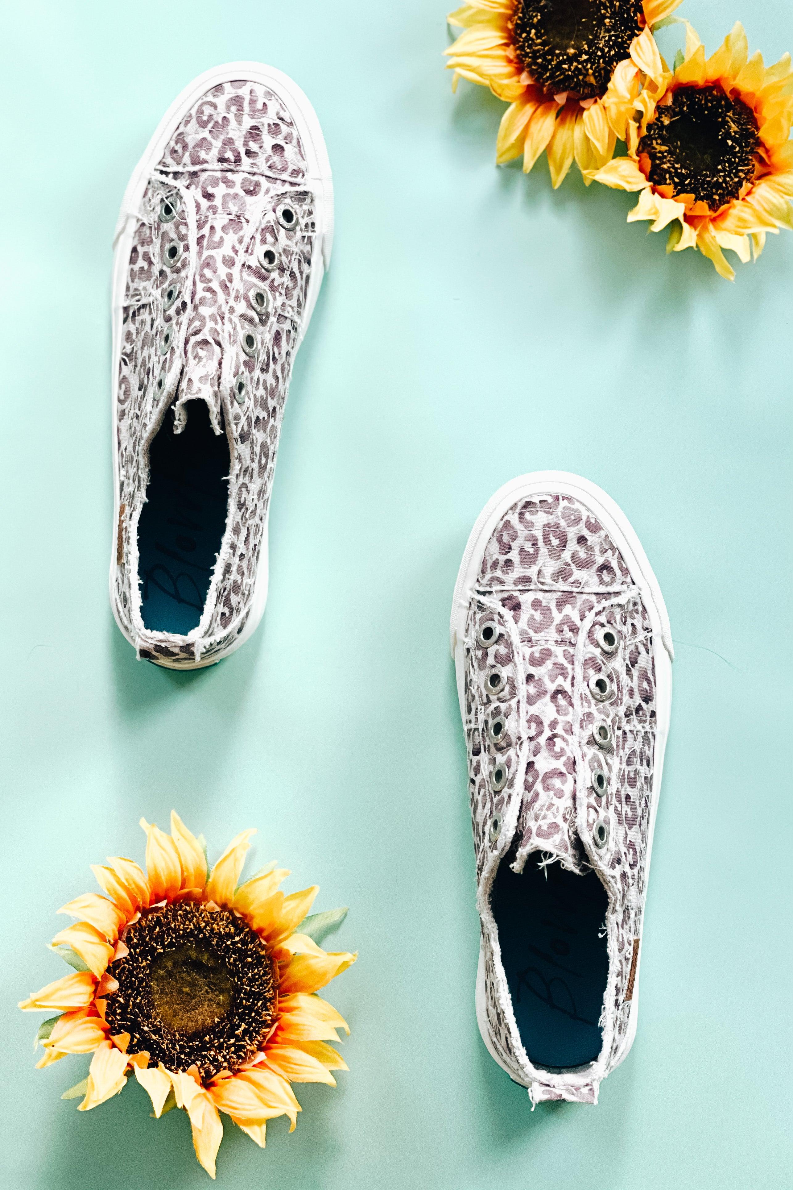 Purple gray water leopard slip-on sneakers displayed flat on pale blue surface with sunflowers nearby