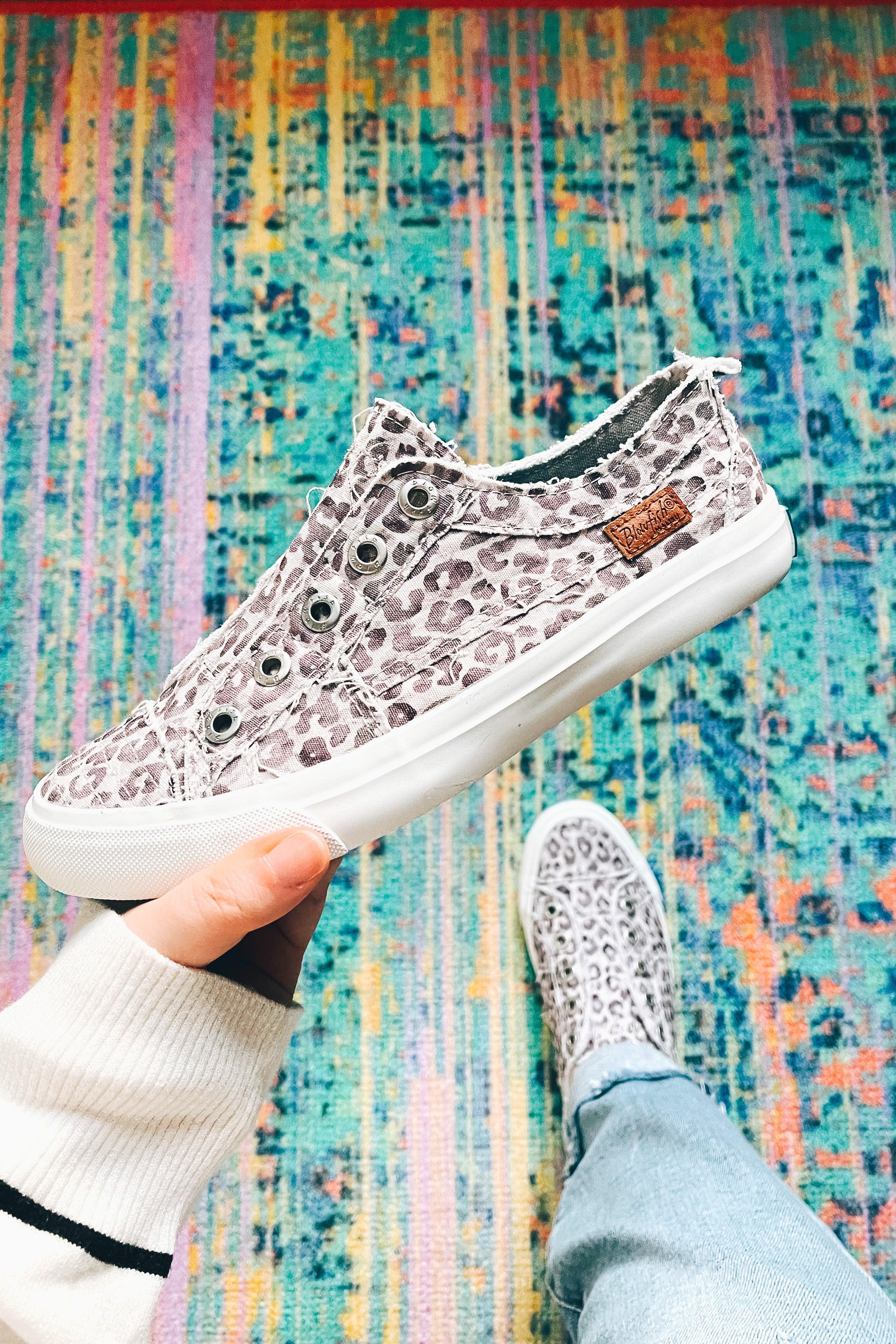 Hand holding purple gray water leopard slip-on sneaker above colorful patterned rug with foot wearing matching shoe