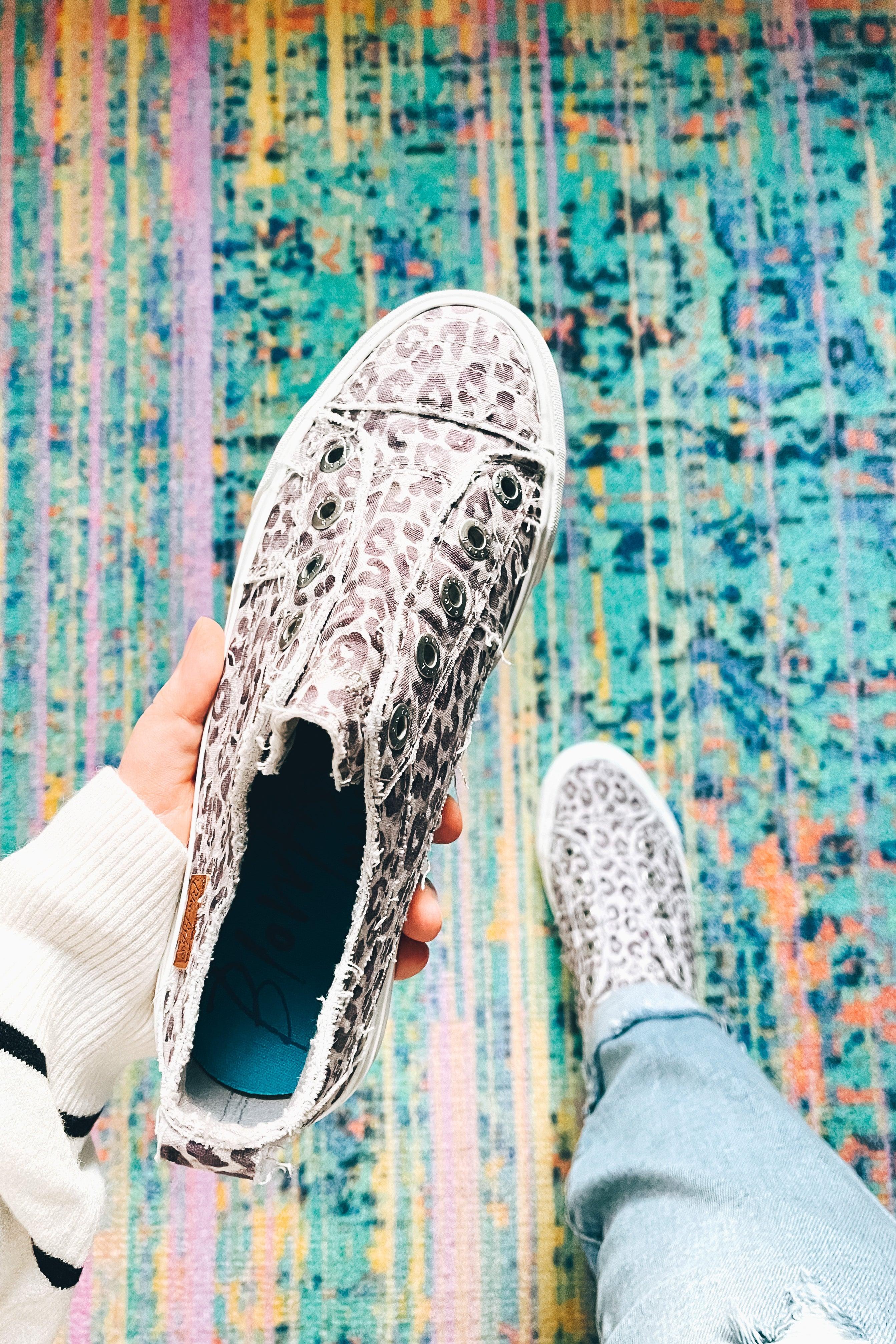 Hand holding purple gray water leopard slip-on sneaker over colorful woven rug with person wearing matching shoe