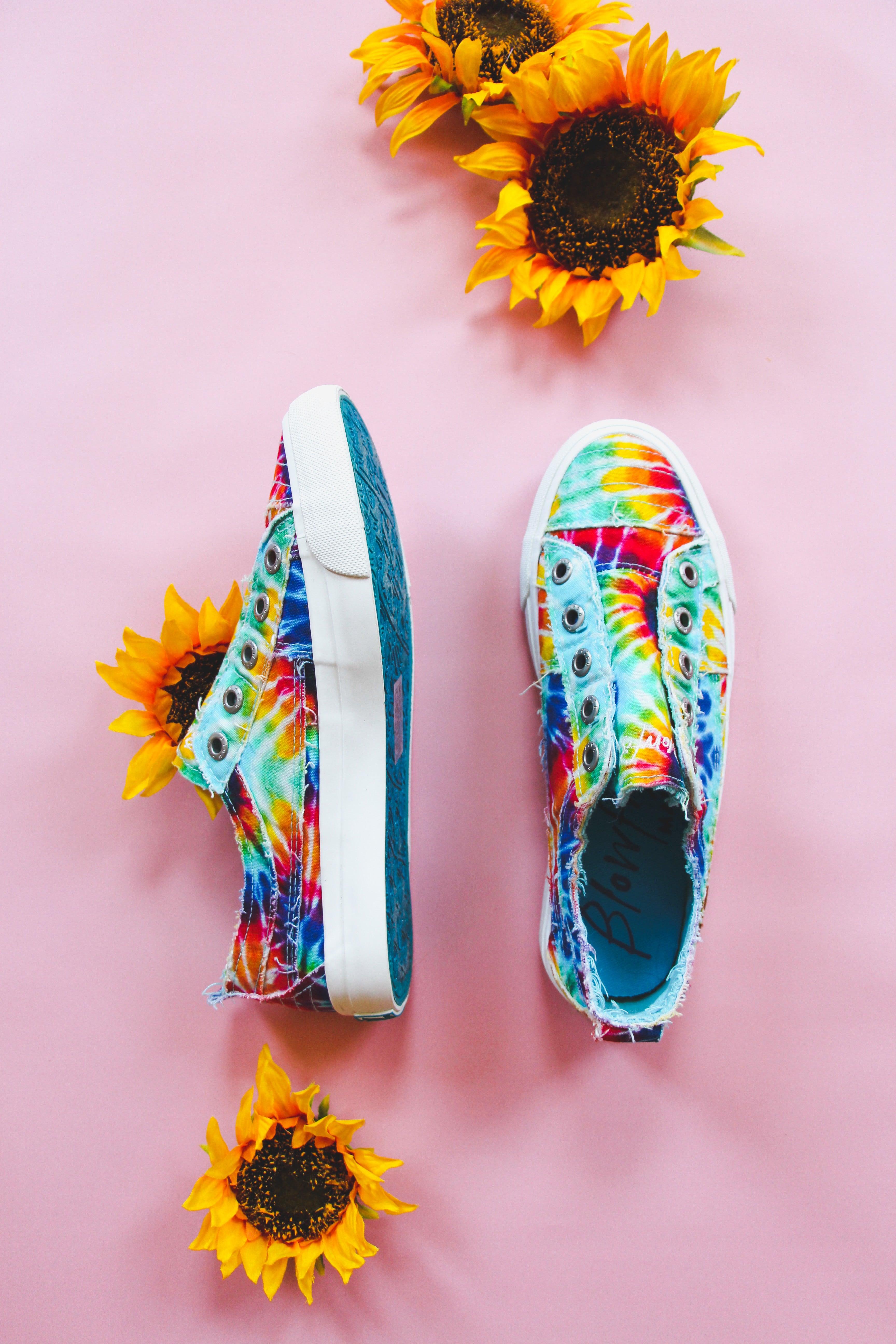 Colorful rainbow tie-dye slip-on canvas sneakers with frayed edges displayed on pink background with sunflowers