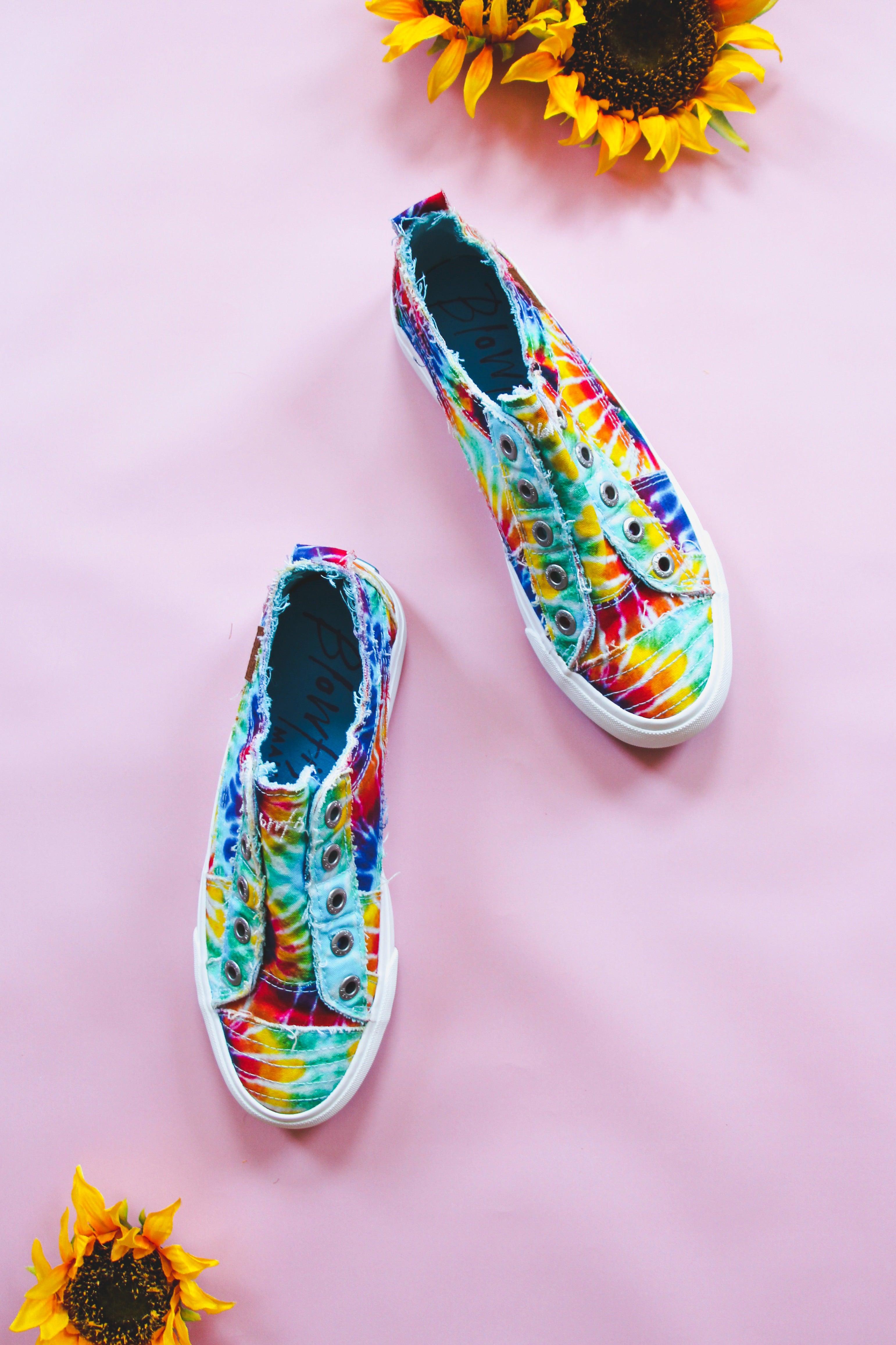 Rainbow tie-dye canvas slip-on sneakers with frayed edges on pink background with two sunflowers nearby