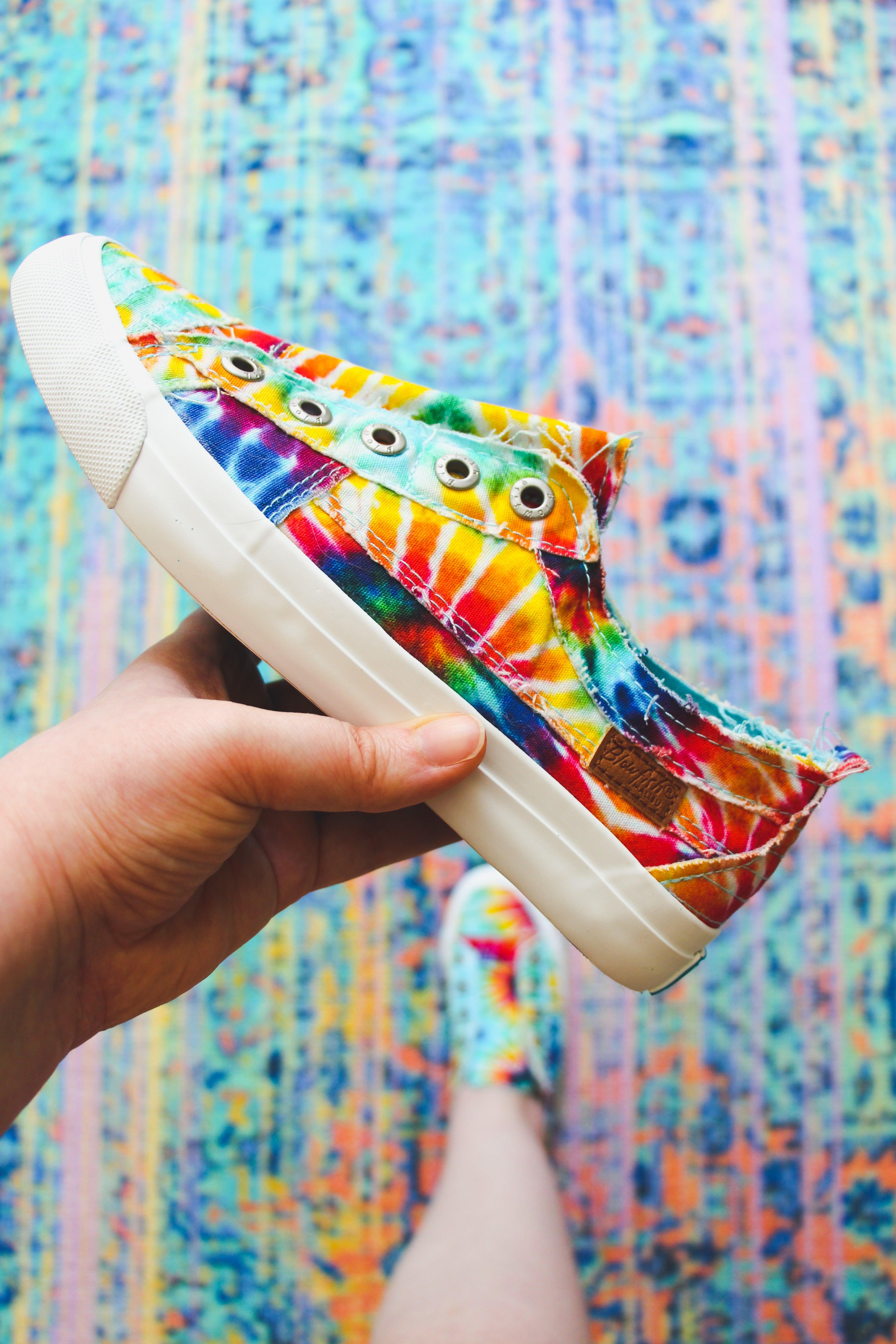 Hand holding colorful tie-dye canvas sneaker with white rubber sole above matching shoe on patterned rug