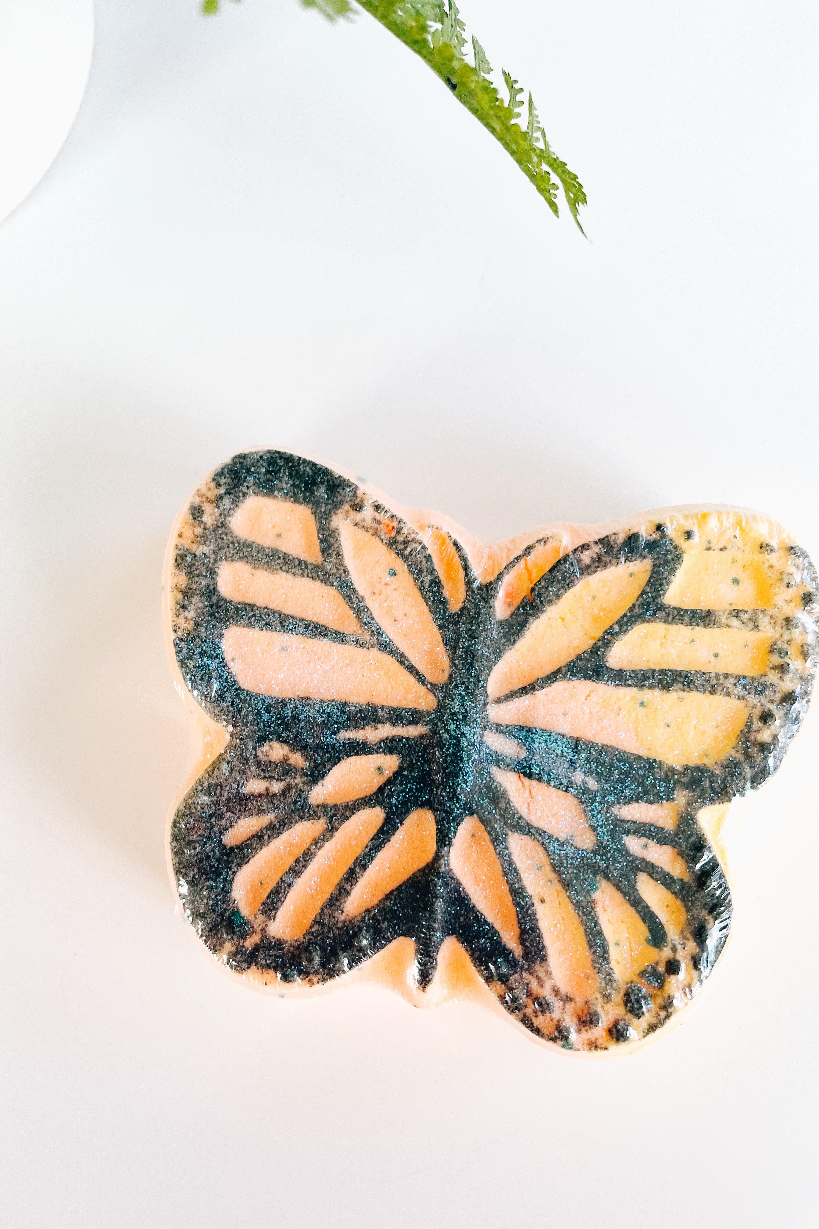 Butterfly-shaped bath bomb with orange, black, and yellow colors on white surface with green fern detail
