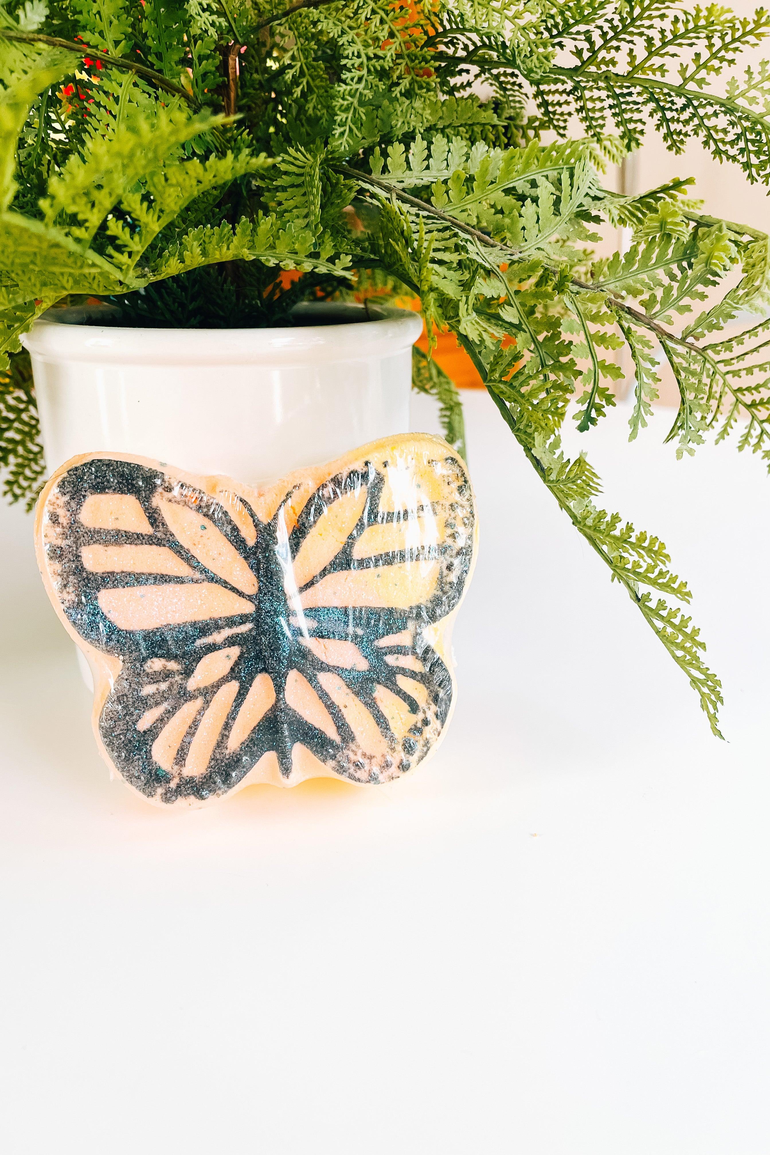 Butterfly-shaped bath bomb with orange and black design placed in front of white planter with green fern leaves