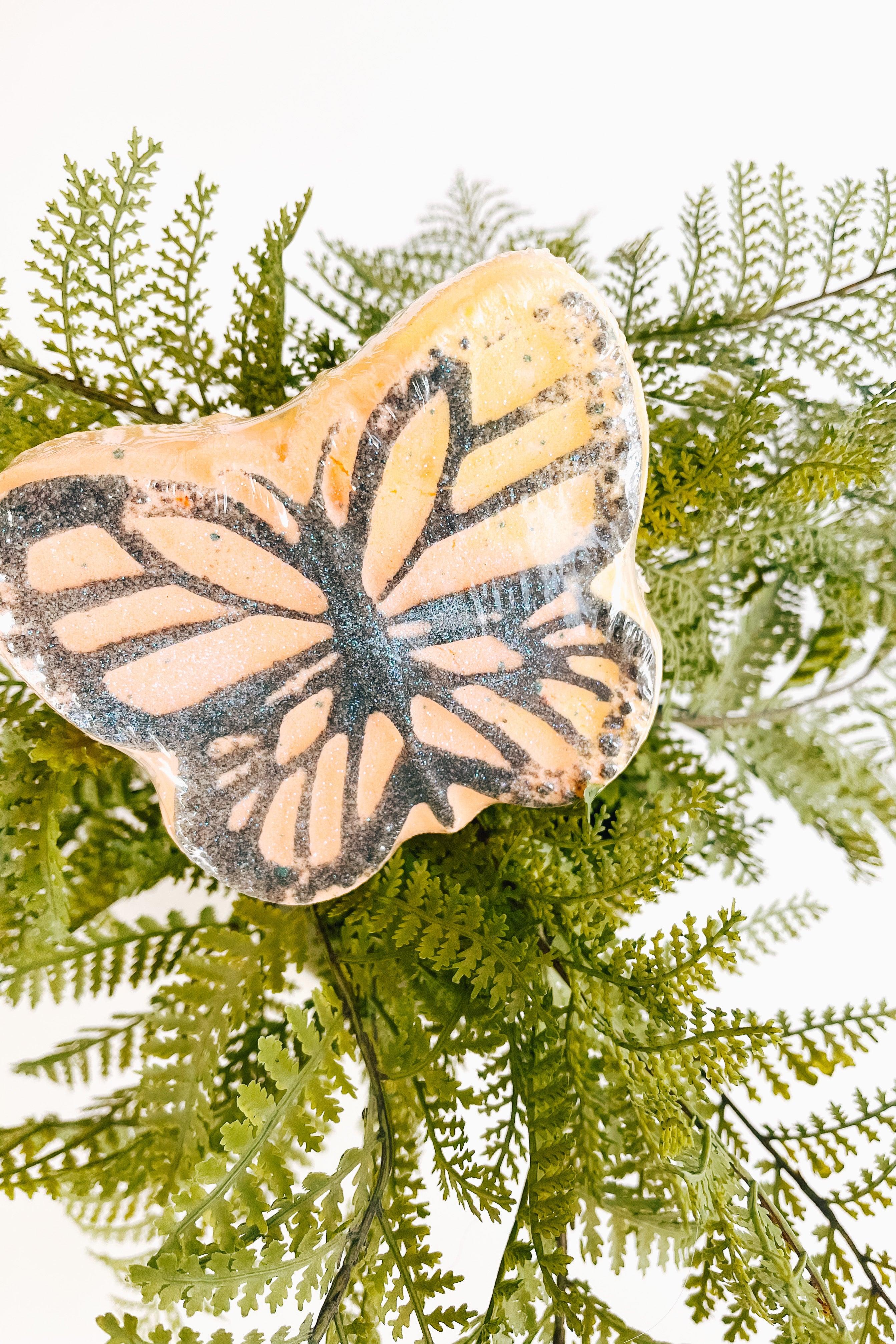 Close-up of butterfly-shaped bath bomb with orange and black pattern resting on green fern leaves against white background