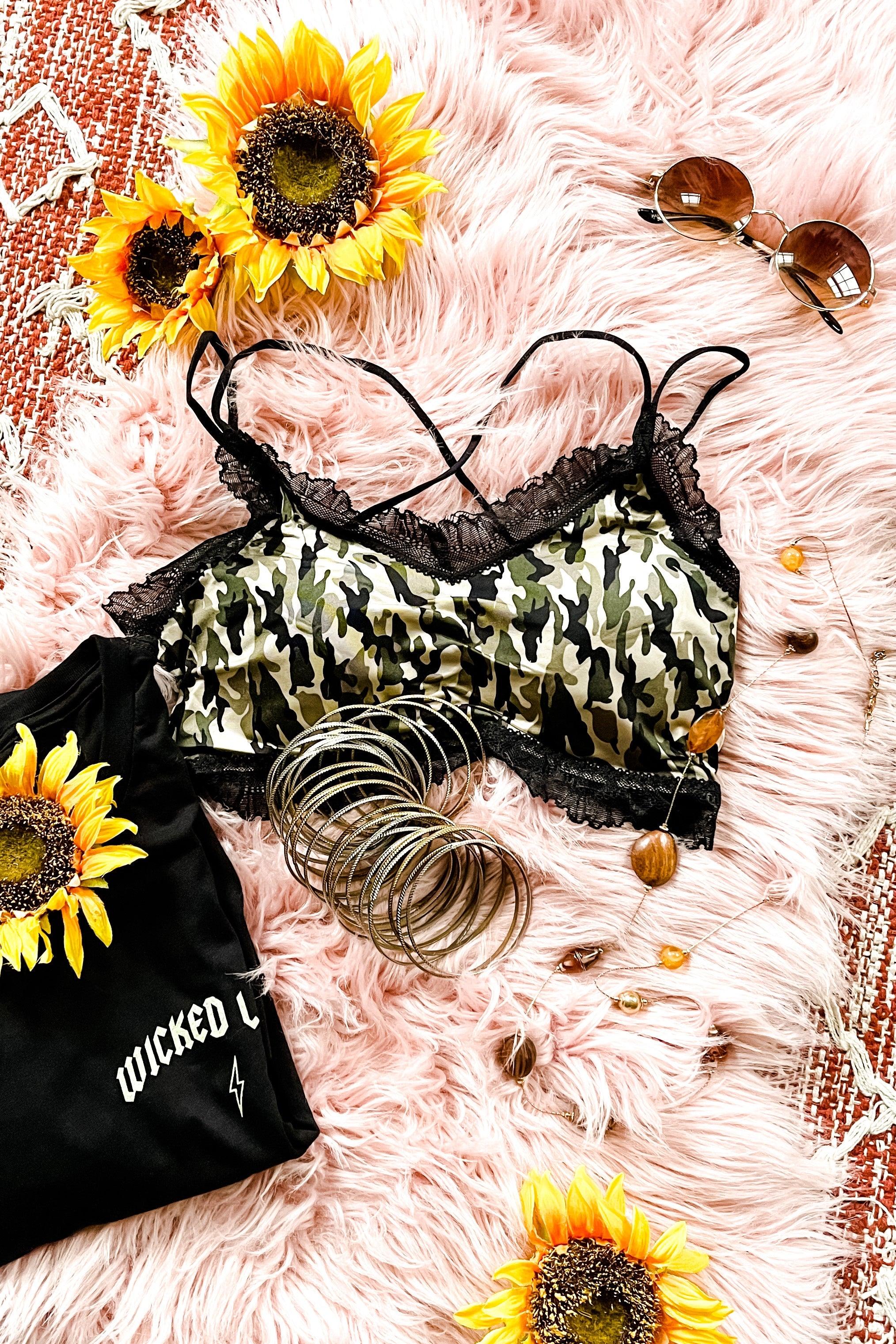 Camo bralette with black lace trim and multiple straps styled with sunflowers, bangles, sunglasses, and black graphic tee on pink rug