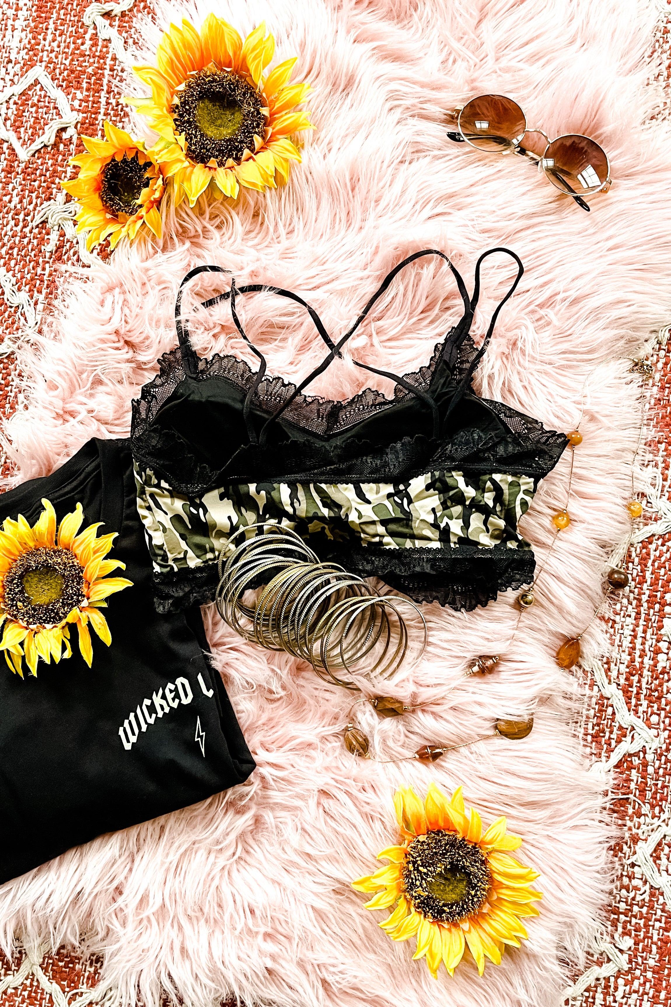Black lace-trimmed camo bralette with multiple straps laid on pink fur rug with sunflowers, bracelets, sunglasses, and black shirt