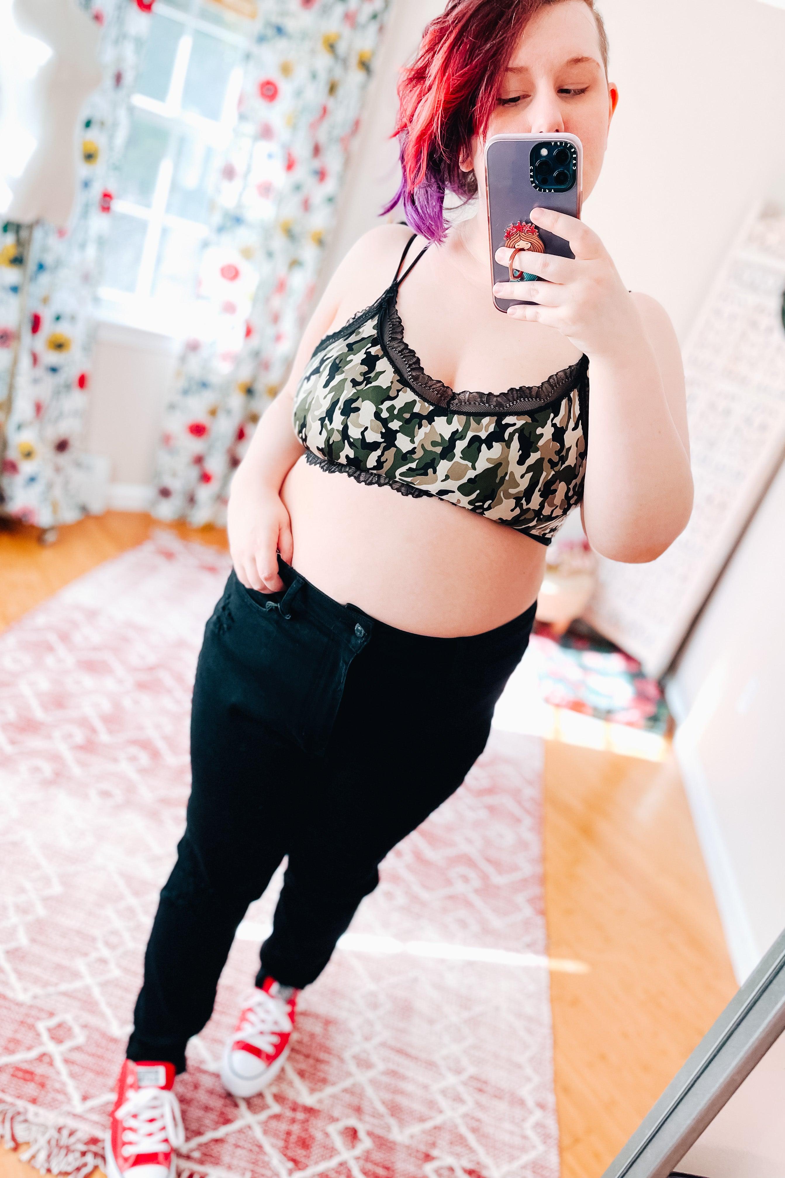 Woman with red and purple hair taking mirror selfie wearing camo bralette with black lace trim and black pants