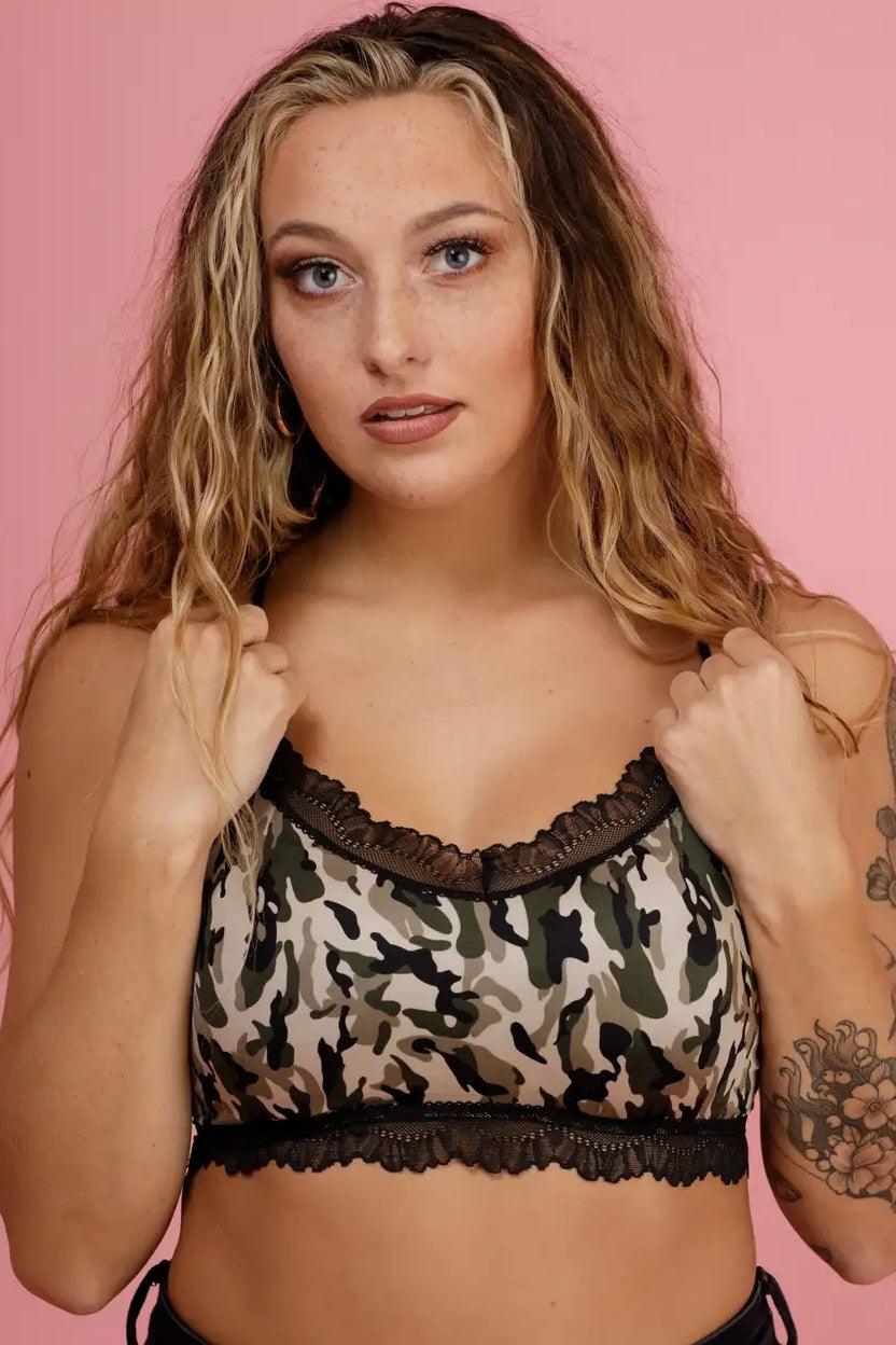 Woman with wavy hair wearing camo bralette with black lace trim standing against pink background