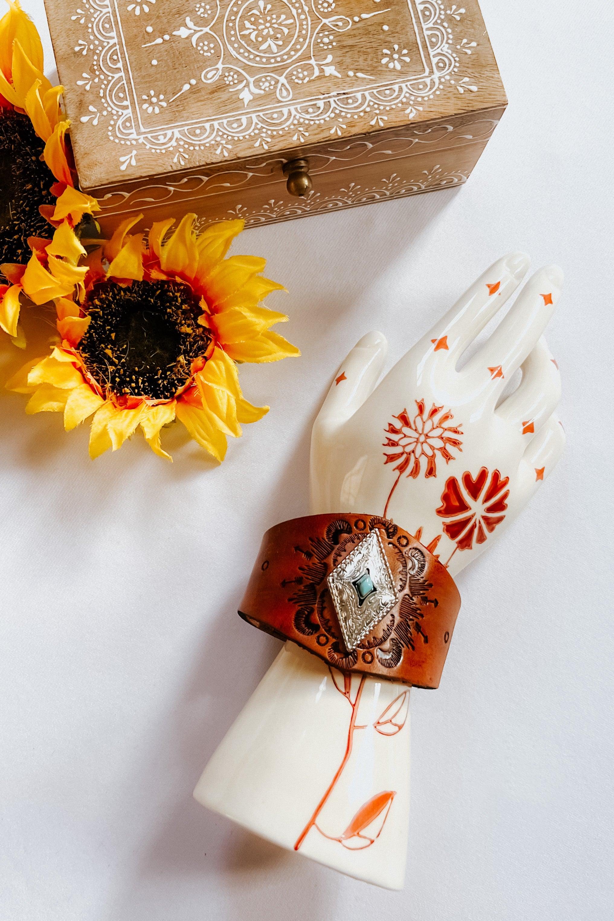 Hand-tooled leather cuff bracelet with silver diamond centerpiece and turquoise stone displayed on painted ceramic hand next to sunflowers and wooden box