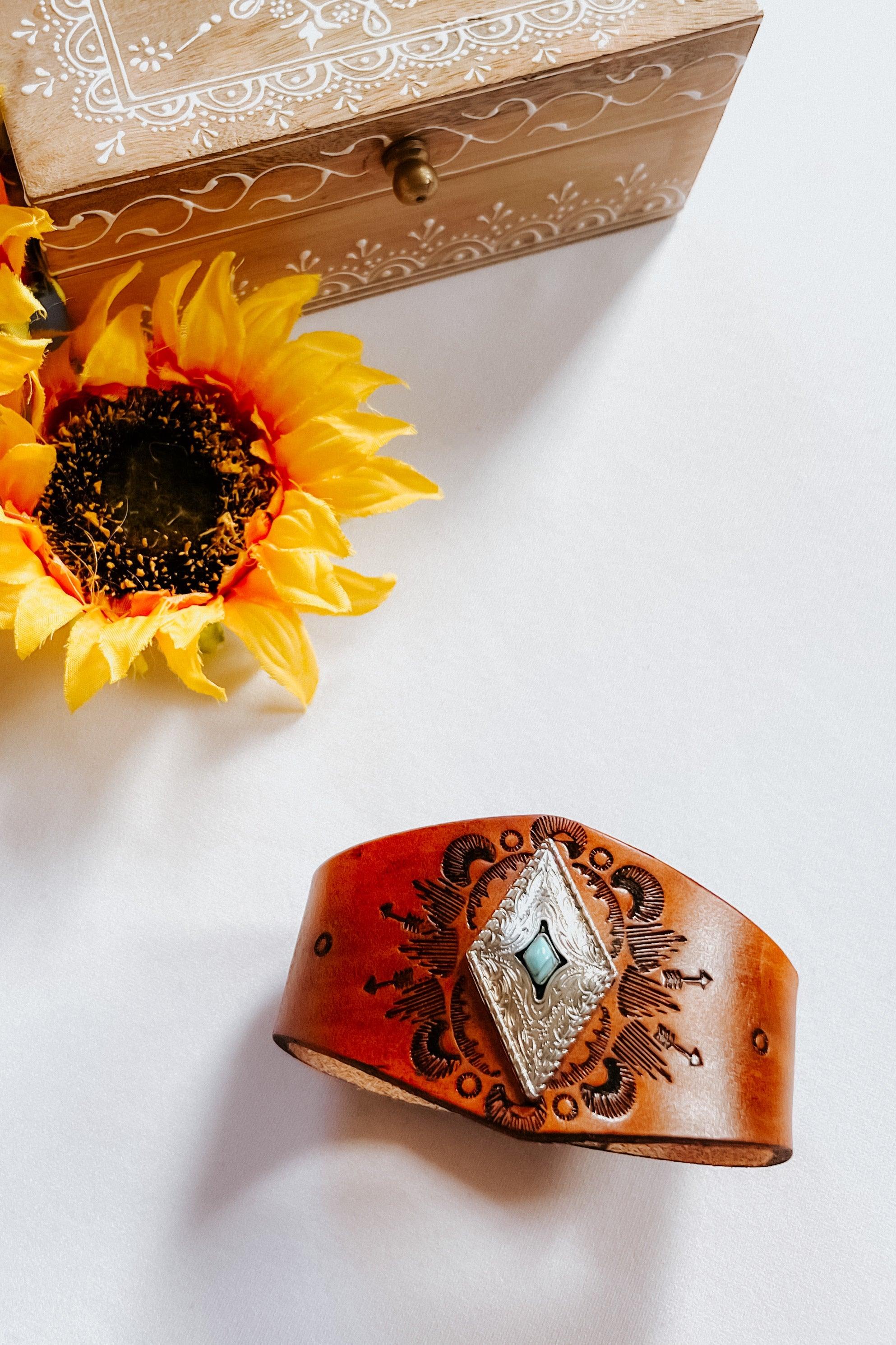 Hand-tooled brown leather cuff bracelet with engraved silver diamond centerpiece and turquoise stone on white surface next to sunflower