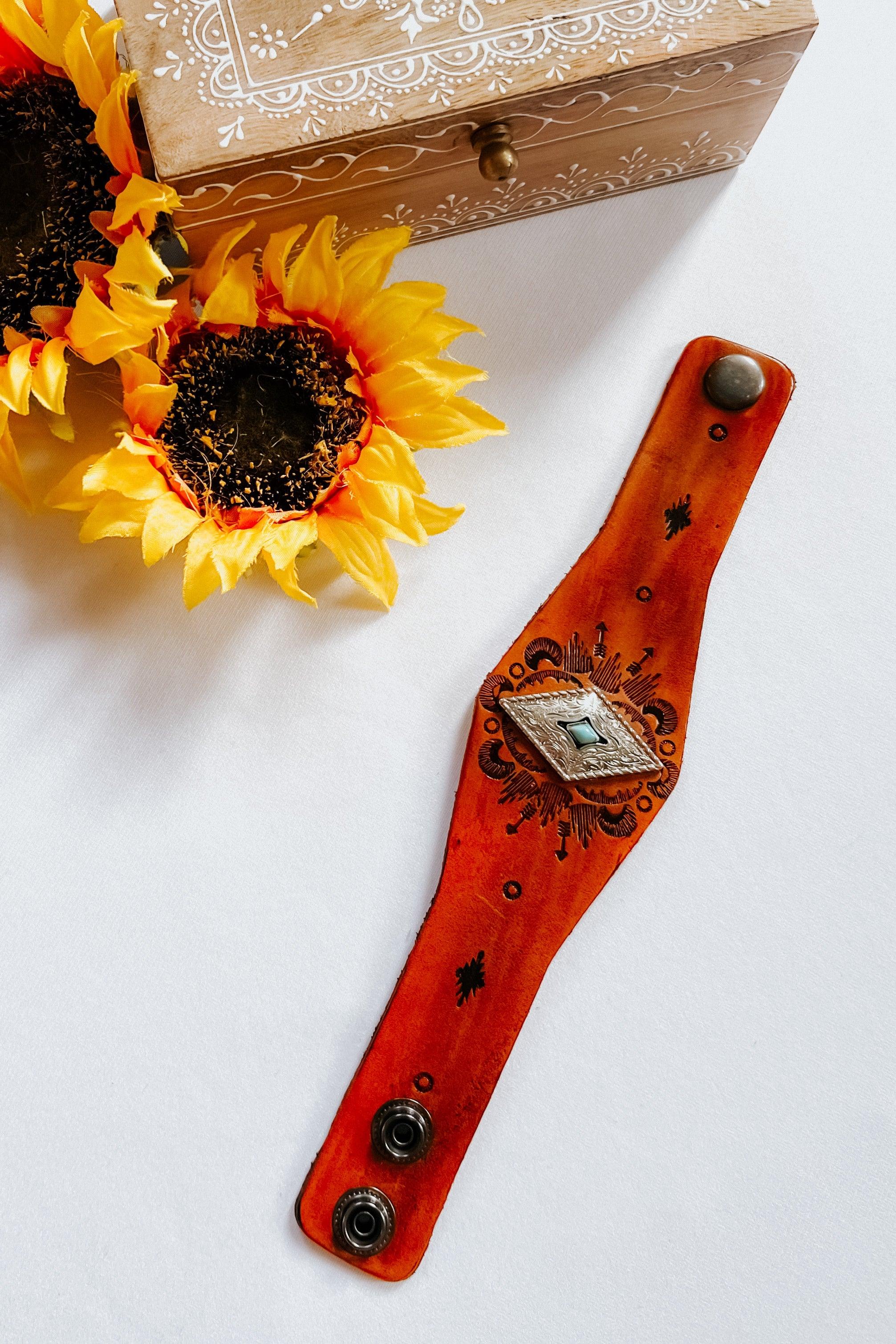 Handcrafted leather cuff bracelet with engraved patterns and central silver diamond accent on white background next to sunflowers and box