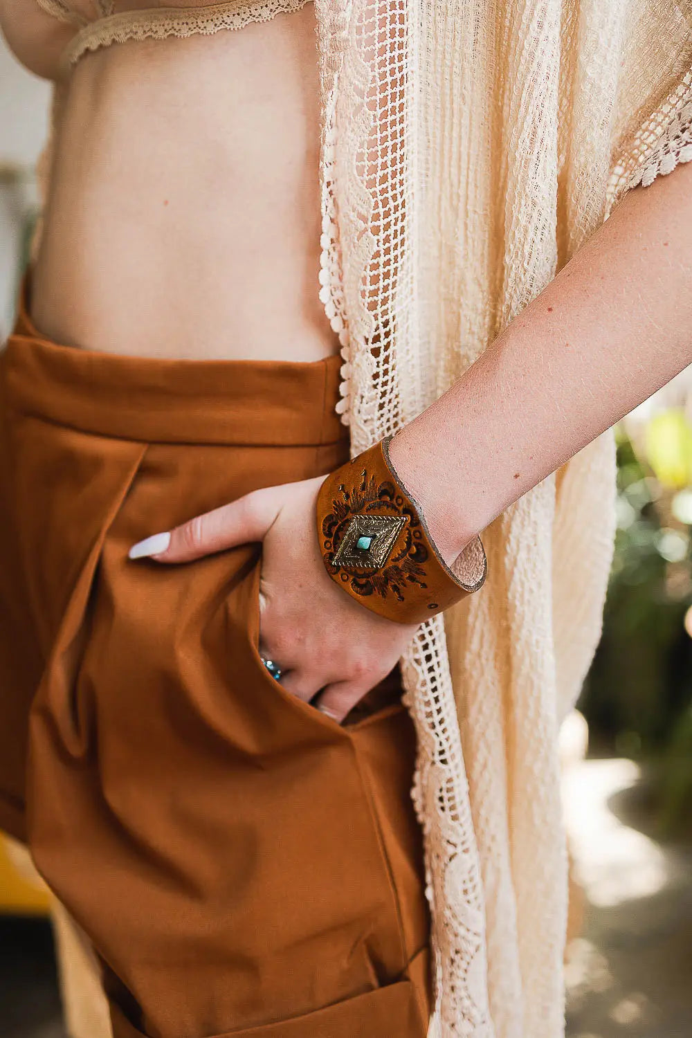 Close-up of wrist wearing a brown leather cuff bracelet with engraved floral design and turquoise stone center
